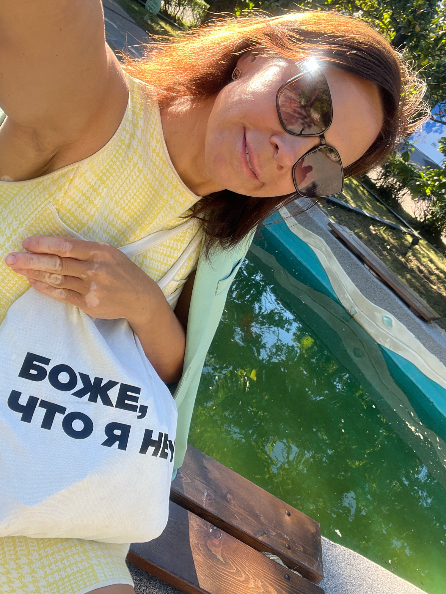 A woman with brown hair and sunglasses takes a selfie outdoors next to a pool. She is wearing a yellow and white patterned dress and a light blue jacket. She holds a white tote bag with black Russian text that reads "БОЖЕ, ЧТО Я НЕ" which translates to "Oh God, what I am not". The pool is filled with green water. The scene appears to be in a park or public area, possibly in Sochi, Russia, given the presence of trees and sunlight.