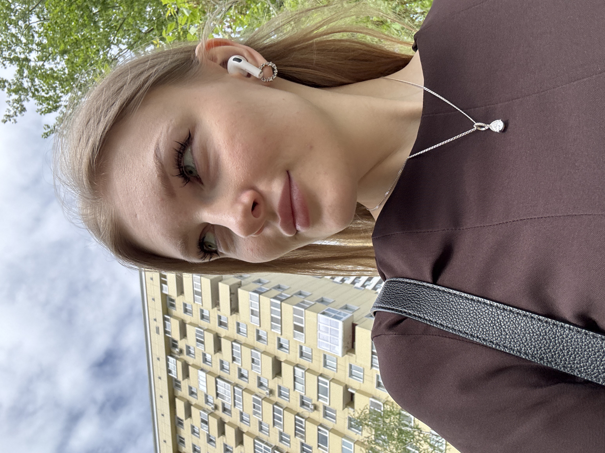 A woman takes a selfie in Perm, Russia. She is wearing a brown shirt, a silver necklace with a teardrop pendant, and an Apple AirPod in her right ear with a rhinestone band around it. A black bag strap is visible across her chest. Behind her is a tall, yellow apartment building with multiple windows, typical of Soviet-era architecture. Green trees are visible in the background along with a cloudy sky.