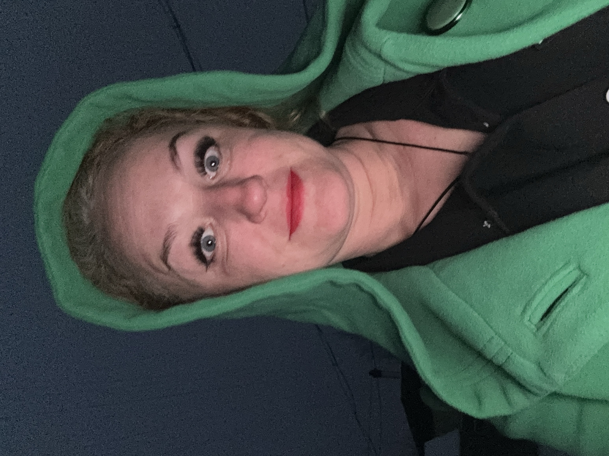 A woman with blonde hair and blue eyes is wearing a green hooded coat and black shirt. She has red lipstick and is looking at the camera. The background is dark blue. It is not possible to determine if this image was taken in Chaikovsky, Russia, or to provide additional context about the scene.