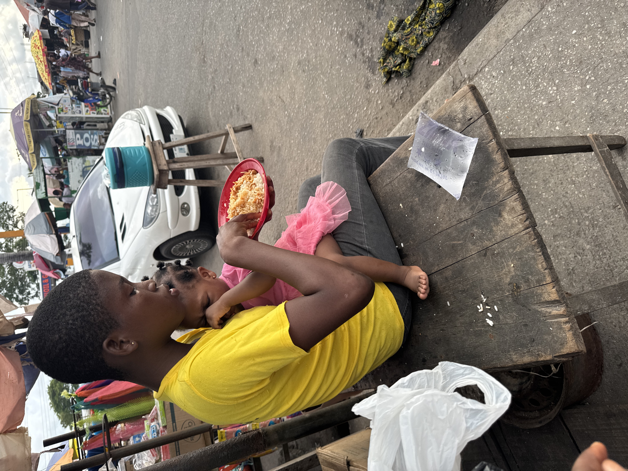 An eye-level shot oriented at an oblique angle shows a young person in a yellow t-shirt and grey jeans sitting on a wooden cart in the city of Badagry, Nigeria. They are holding a young child wearing a pink dress in their lap. The child is angled towards the young person and appears to be eating from a red plate filled with rice. A clear plastic bag filled with water is on the cart, as well as a white plastic bag. The background includes a white car, various vendors and pedestrians along the street, and buildings and trees in the distance.