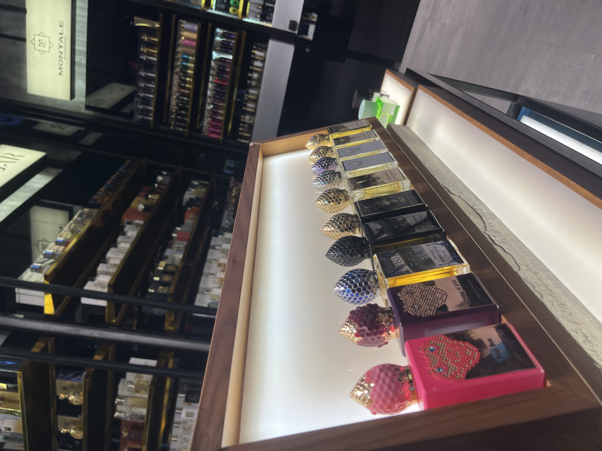 A retail display case containing a variety of perfumes, with the bottles arranged in a neat row, is situated in a shop environment. The perfume bottles have different colors and designs. The foreground is a display case with a wooden frame. The background shows shelves filled with more perfume bottles. Based on the signage visible in the background, the scene may be in Sofia, Bulgaria, as the 'Montale' perfume brand has a store there. The perspective of the image is slightly off-center.