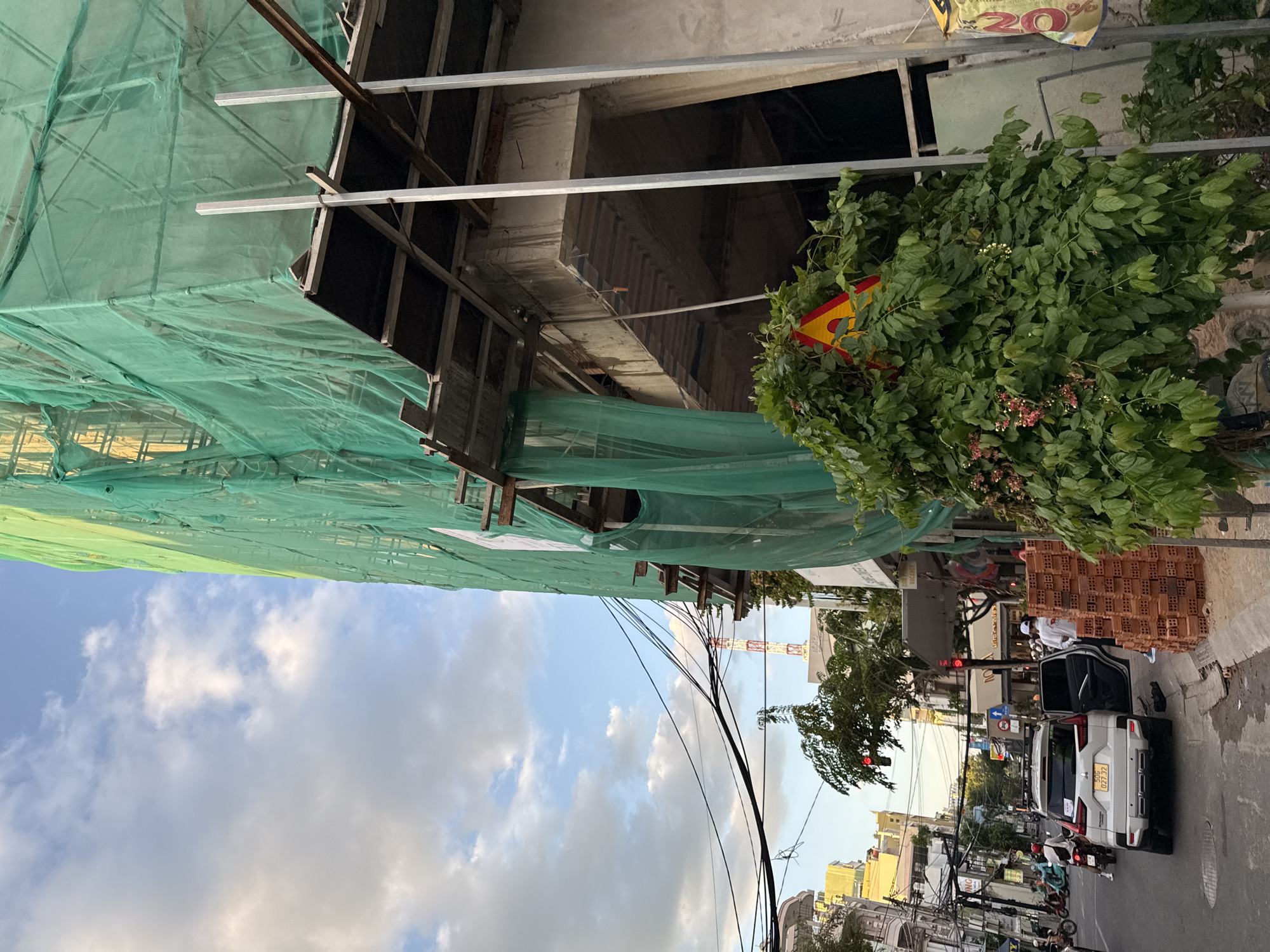 The image depicts a street scene in Nha Trang, Vietnam, with a building under construction prominently featured. The building is covered in green netting, obscuring most of its structure. Scaffolding and construction materials are visible. In the foreground, a dense green bush with small pink flowers partially obstructs a yellow and red triangular warning sign. To the left, a white SUV is parked on the street, with its front passenger door open. Two individuals in white shirts and hats stand beside the SUV. A stack of red bricks is piled on the sidewalk next to them. The street is lined with other buildings, some appearing older and multi-story, with balconies and awnings. Electrical wires are strung overhead, characteristic of urban environments. Further down the street, traffic signals and other vehicles, including motorcycles, are visible. The sky is partly cloudy with a mix of blue sky and white clouds. The overall scene suggests an active urban environment with ongoing construction.