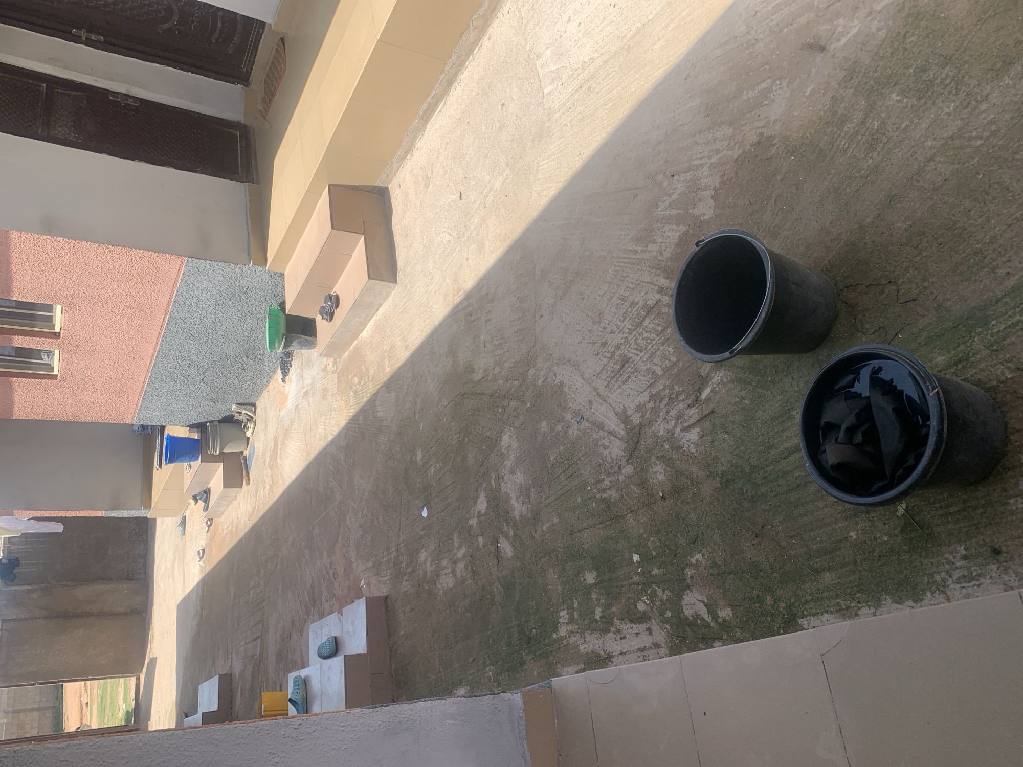 The image shows an outdoor walkway in Jauro Yinu, Nigeria. The walkway is paved with concrete and shows signs of wear and moss growth. To the left, there are concrete steps leading up to a building. On these steps, there are two pairs of blue flip-flops and a yellow bucket. Further down the walkway, there are more concrete steps leading to a building with a pink and grey textured facade. On these steps, there is a blue bucket, a grey bucket, and a pair of sandals. Two dark-colored buckets are visible on the ground further down the walkway; one is empty and the other contains dark fabric and water. The area is partially shaded, suggesting it is daytime. The buildings appear to be residential or small commercial structures.