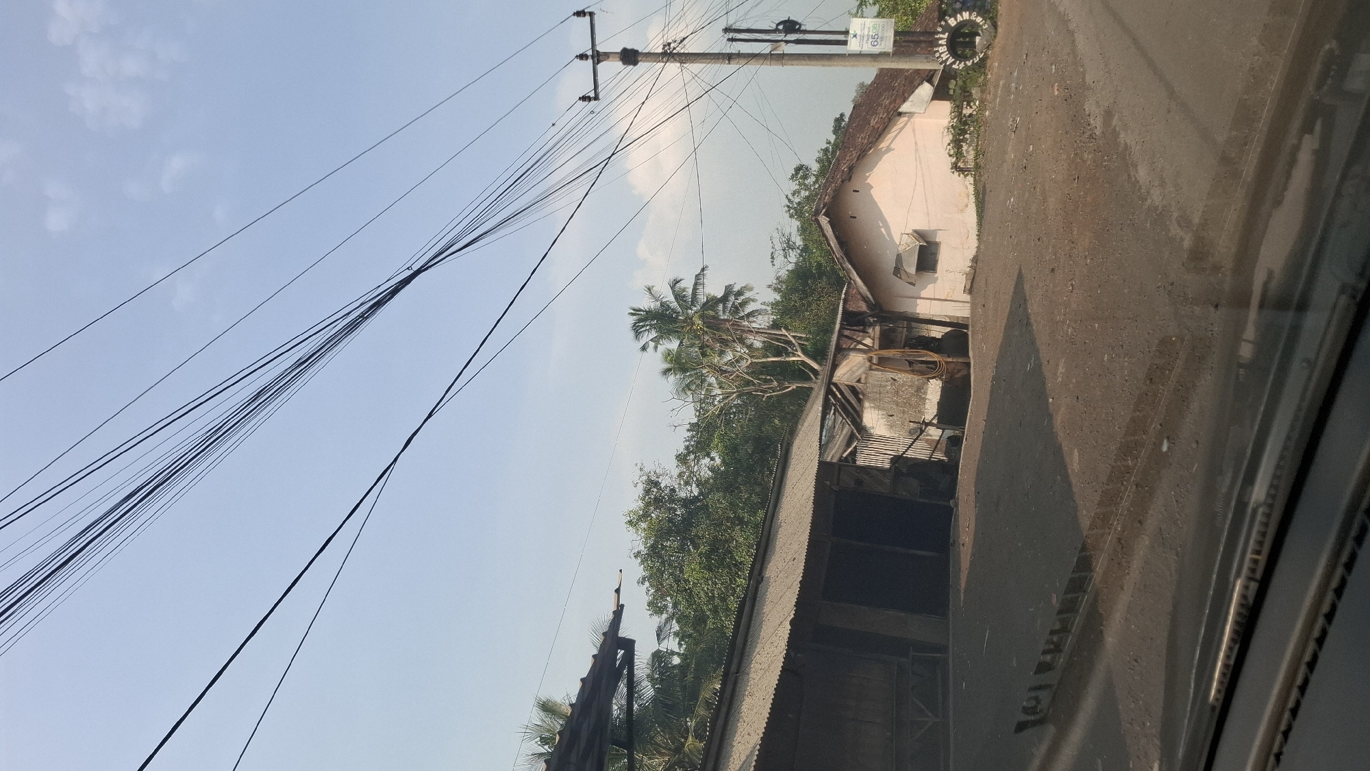 The image depicts a street scene in Sirnagalih, Indonesia, characterized by a dusty road in the foreground, reflecting the bright sunlight. To the left, a dilapidated structure with a corrugated metal roof stands prominently, appearing aged and weathered. Adjacent to it, another building with a similar roof is visible. Further to the right, a light-colored house with a brown tiled roof is situated, featuring a small window. Overhead, a dense network of power lines crisscrosses the sky, supported by a utility pole on the right side of the frame. Beyond the buildings, lush green trees and palm trees dominate the background, suggesting a semi-rural or suburban environment. The overall impression is one of a modest, possibly developing, residential area under a clear blue sky.