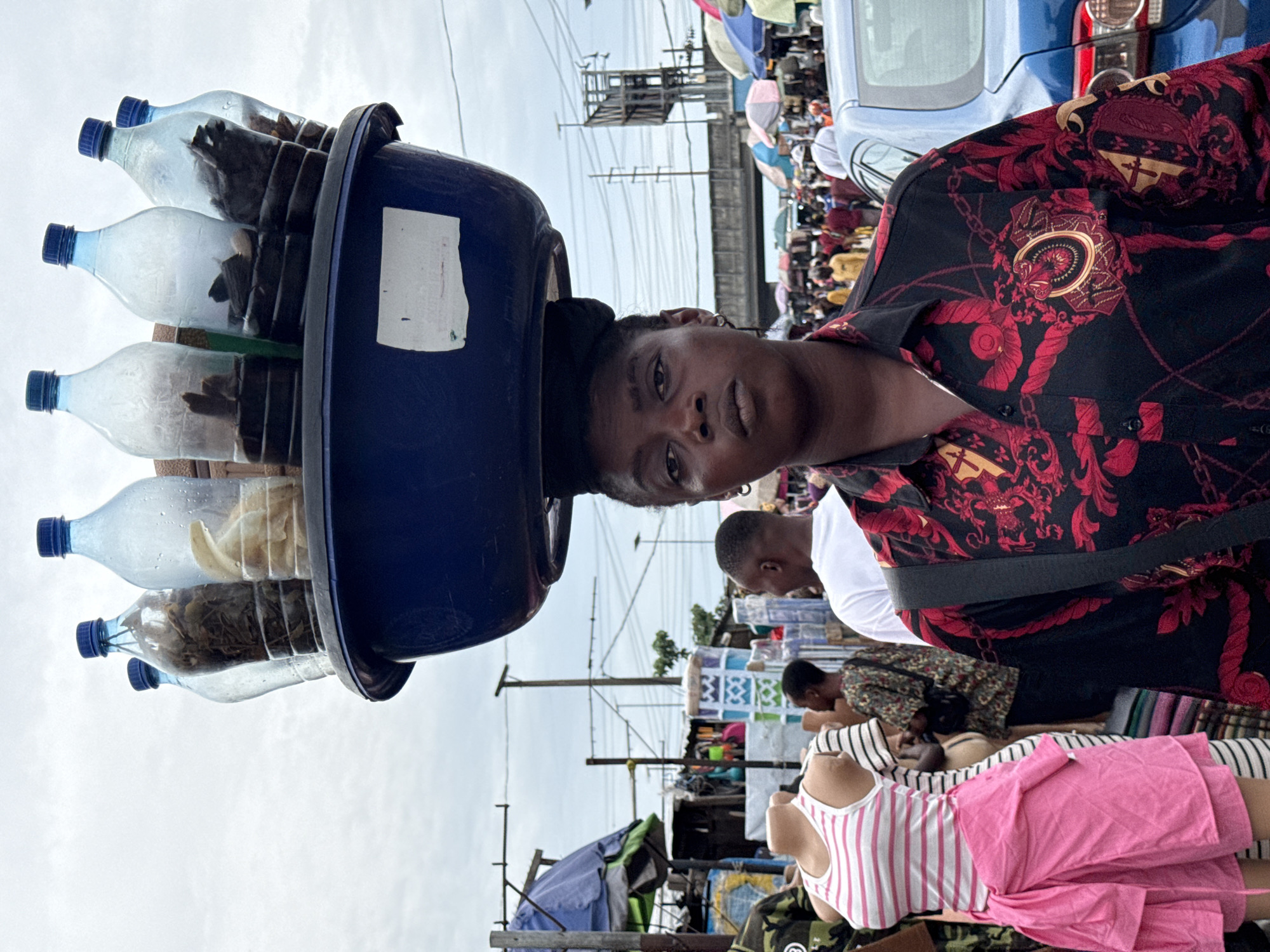 A woman in Ado, Nigeria, stands among a bustling market scene. She balances a blue basin on her head. Inside the basin, plastic bottles are arranged horizontally, each filled with different dry goods. She wears a patterned black and red shirt with a shoulder bag. Around her, various stalls and individuals populate the background, including a mannequin dressed in pink and white, a man in a white shirt, and an overhead view of market umbrellas and power lines. The sky is overcast.