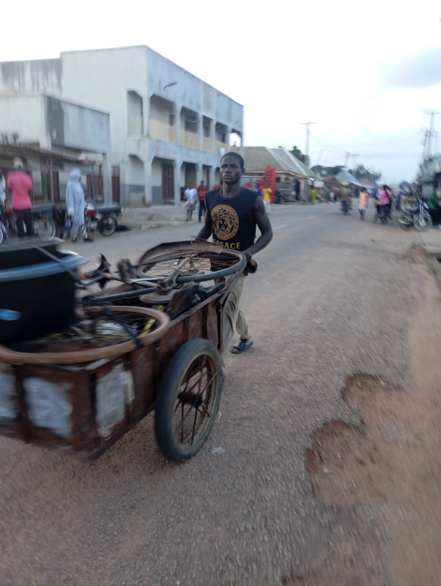 A full-shot, eye-level, outdoor, medium-long shot captures a man in Shendam, Nigeria, pushing a heavily loaded, rusted, two-wheeled cart. The man, facing forward and looking directly at the viewer, wears a black tank top with a gold Versace logo and beige pants. He is wearing dark sandals. The cart, made of what appears to be scrap metal, is filled with bicycle parts, including wheels, frames, and tires. The background shows a street scene with a light-colored, two-story building on the left, typical of urban Nigerian architecture. Other people and motorcycles are visible in the distance, suggesting a busy market or thoroughfare. The road surface is asphalt with patches of dirt and a few visible potholes. The sky is overcast with a hint of blue, indicating possibly early morning or late afternoon. The overall image conveys a sense of daily life and commerce in the city.