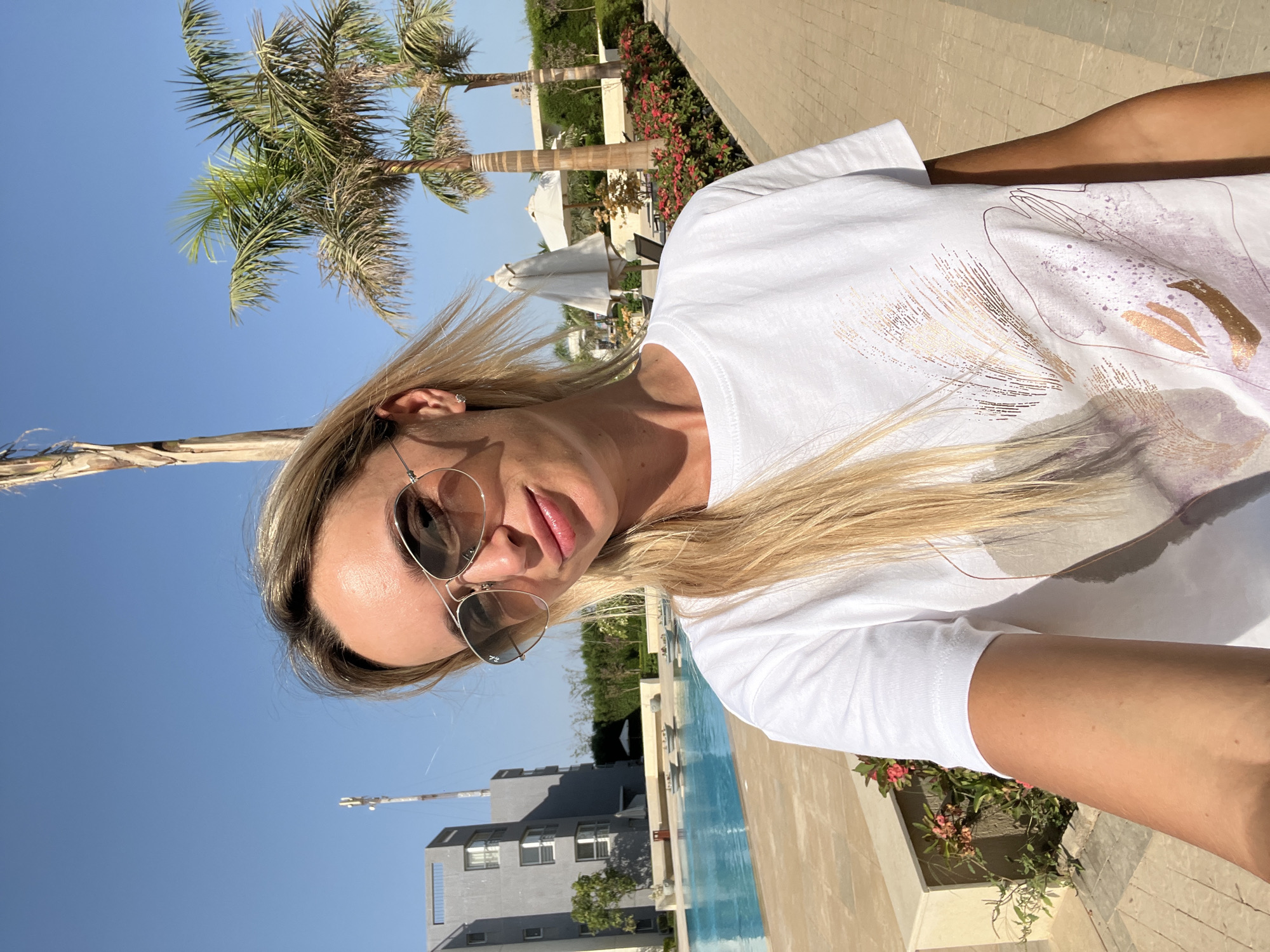 A woman with blonde hair and sunglasses smiles at the camera in a selfie. She is wearing a white t-shirt with a patterned design. She is standing outdoors, with a swimming pool and palm trees visible in the background. A modern building is also visible in the distance. The scene appears to be a resort or hotel in a warm climate.