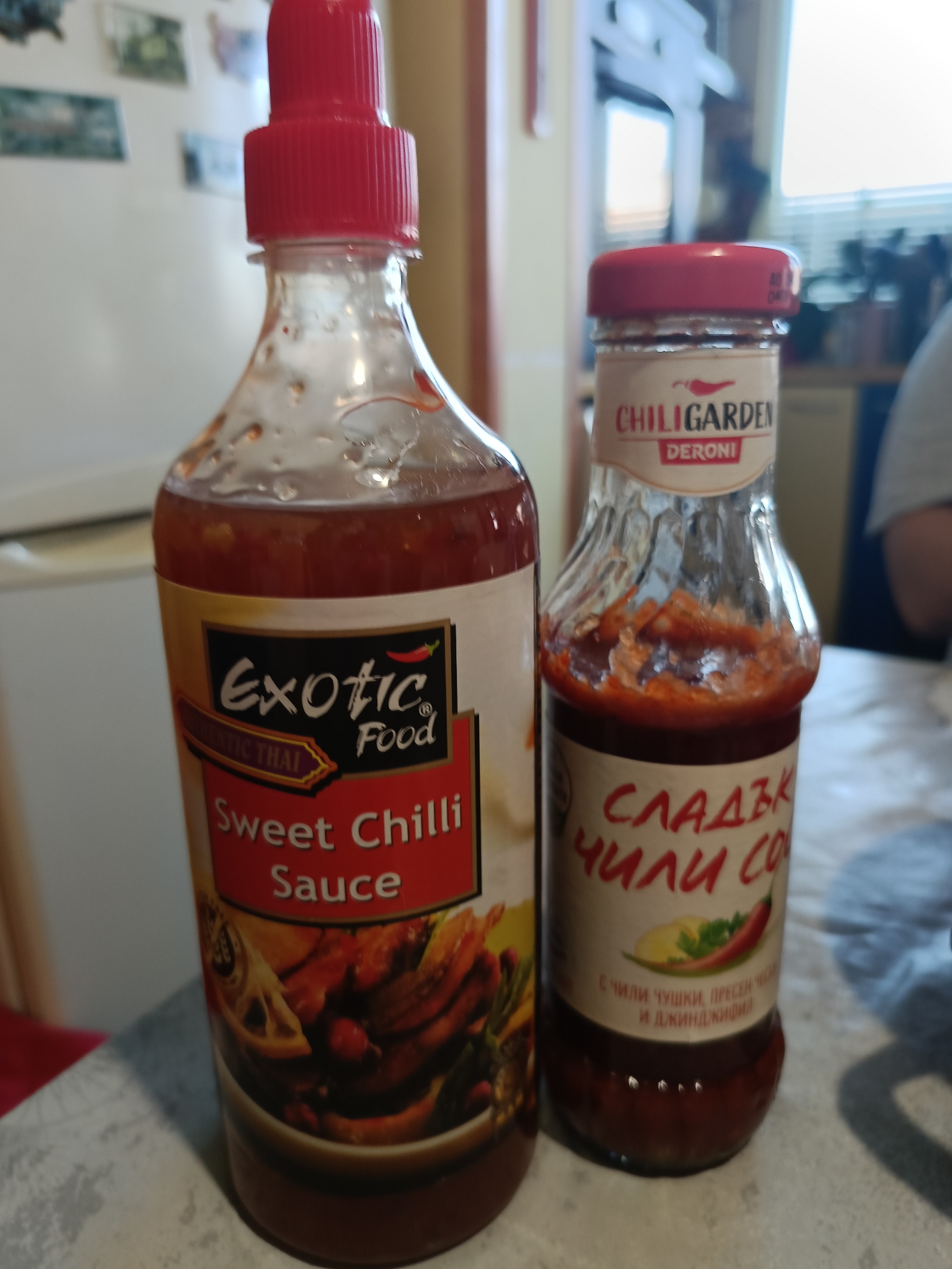 Two bottles of chili sauce are displayed on a table. The bottle on the left is a large, clear glass bottle with a red cap and a label that reads "Exotic Food Sweet Chilli Sauce Authentic Thai". The bottle on the right is smaller, also clear glass with a red cap and a label that reads "CHILIGARDEN DERONI". The sauce in this bottle is a vibrant red. The background of the image is blurred, but appears to be an indoor setting, possibly a kitchen. The lighting suggests it is daytime. There is no indication of the city of Varna, Bulgaria, or any specific location within it.