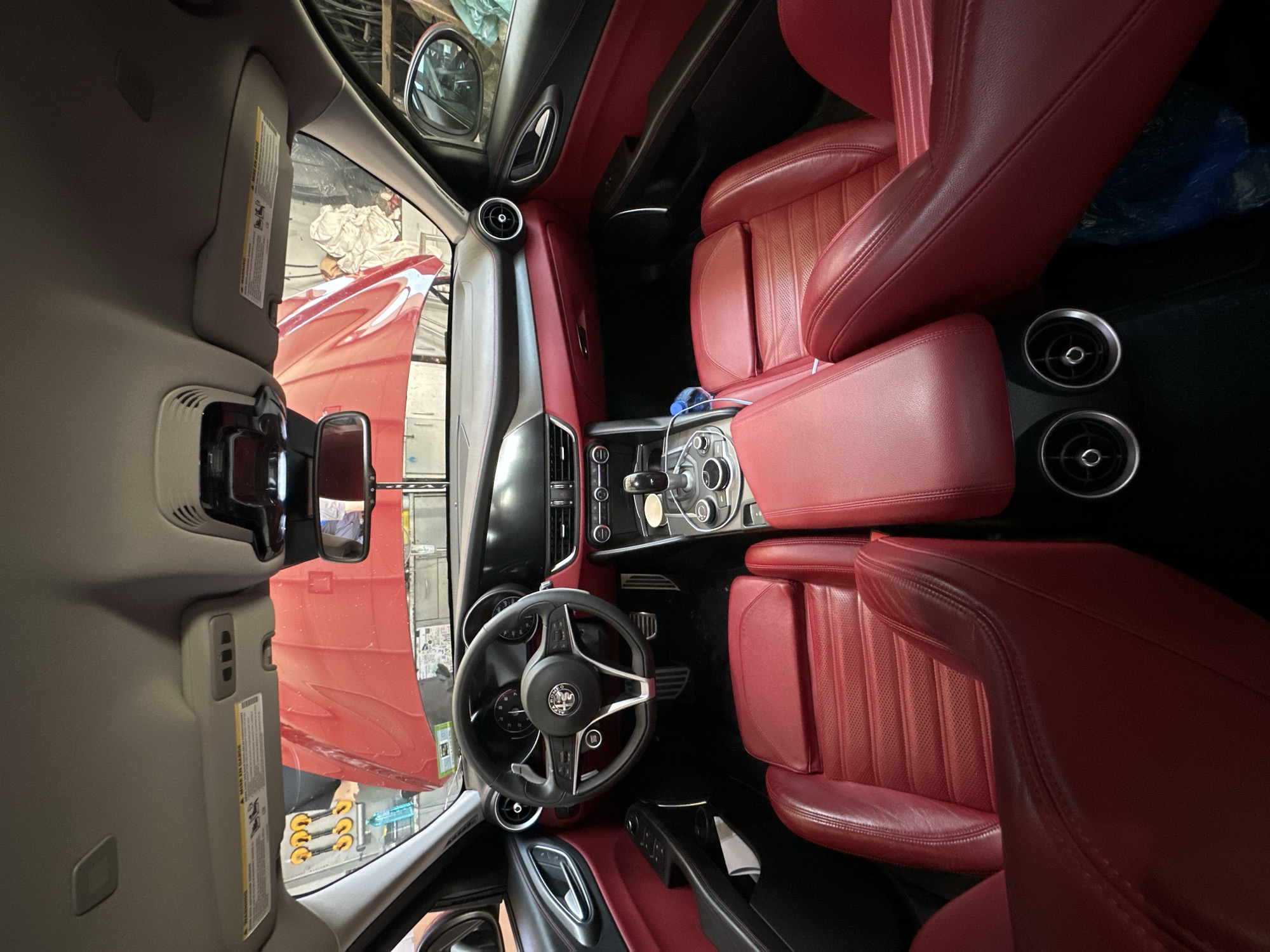 This is an interior shot of a red car, viewed from the back seat looking forward. The car's hood is open, revealing a red engine bay. The interior features red leather seats, a black steering wheel with a silver emblem, and a black dashboard. Two circular air vents are visible on the center console. The car appears to be parked indoors, possibly in a garage or workshop, as indicated by the tools and equipment visible through the windshield. The lighting suggests it is daytime.