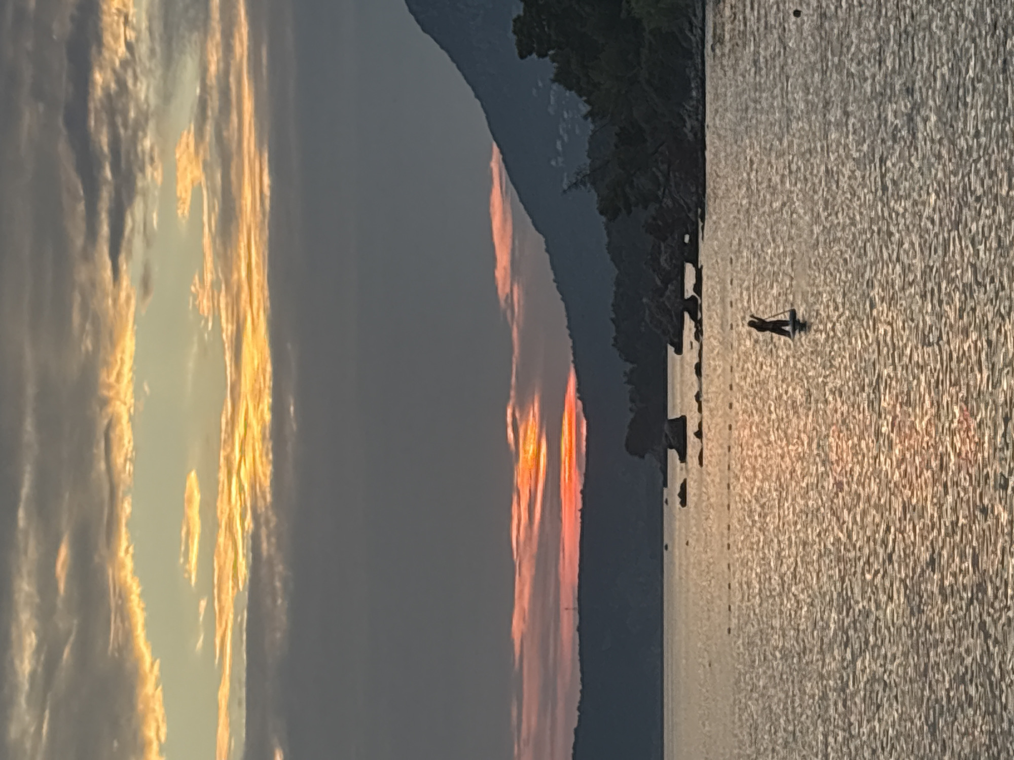 The image captures a serene sunset scene over the Adriatic Sea, likely near Bar, Montenegro. The sky is a canvas of warm colors, with streaks of orange and yellow peeking through dramatic grey clouds. The sun's rays cast a golden shimmer across the rippling water, creating a tranquil atmosphere. In the distance, a silhouette of a person on a paddleboard can be seen, enjoying the peaceful evening. The coastline is visible with rolling hills and rocky shores, dotted with lush green trees. The overall impression is one of natural beauty and quietude.