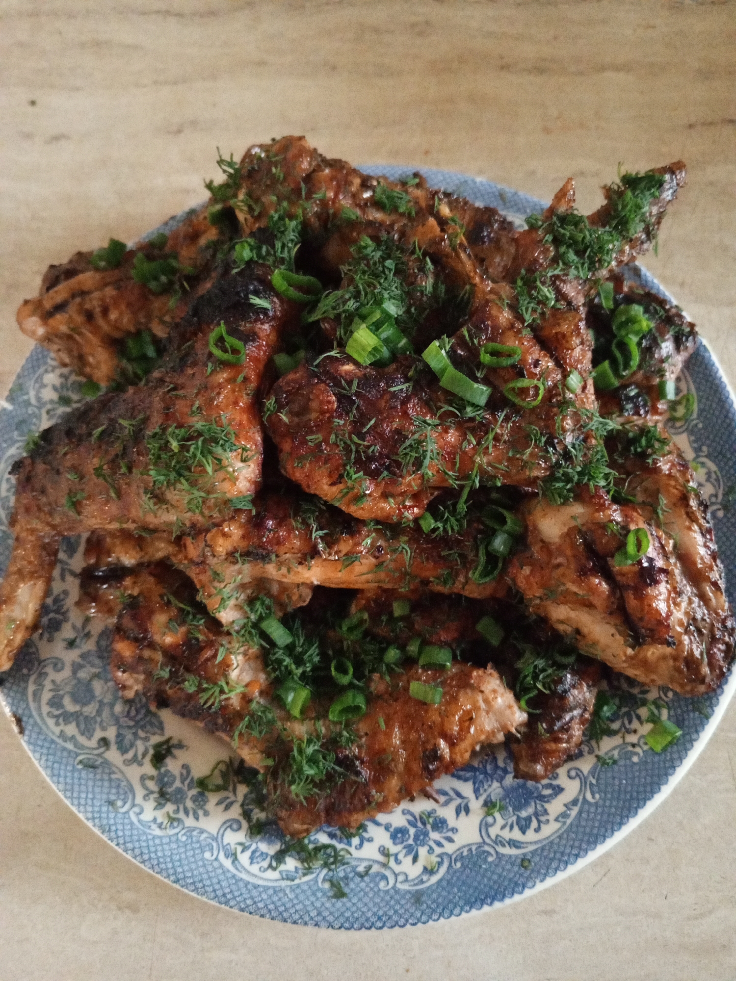 A plate of grilled chicken wings is presented, garnished with chopped green onions and dill. The wings are a rich brown color with visible grill marks, suggesting they have been cooked over a flame. The plate itself has a blue and white floral pattern. The background is a neutral, textured surface that appears to be a countertop. No people are visible in the image, and no specific location in Buraly, Russia can be determined from the provided visual information.