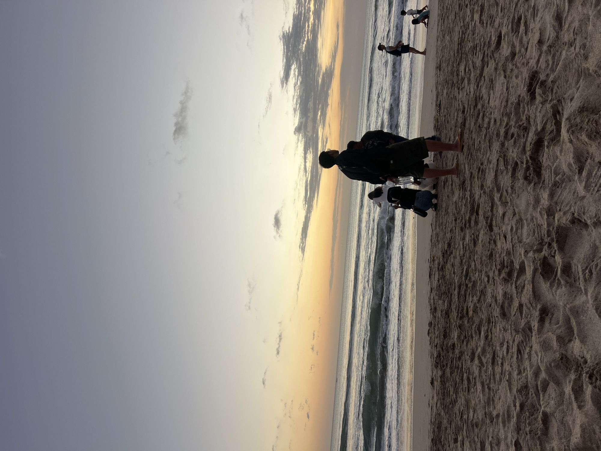 The image depicts a beach scene in Kuta, Indonesia, likely during either sunrise or sunset due to the gradient of the sky from pale blue to soft orange. In the foreground, the sandy beach is textured with footprints and undulations. A few people are scattered across the sand. The middle ground shows the ocean with moderate waves breaking on the shore. Several individuals are visible on the beach, some standing and looking out at the sea, while others are seated or in the process of moving. One person in the center foreground is standing and facing away from the camera, appearing to observe the horizon. Further in the distance, more figures can be seen near the water's edge and further down the beach. The sky above is a soft, hazy expanse with scattered, wispy clouds.