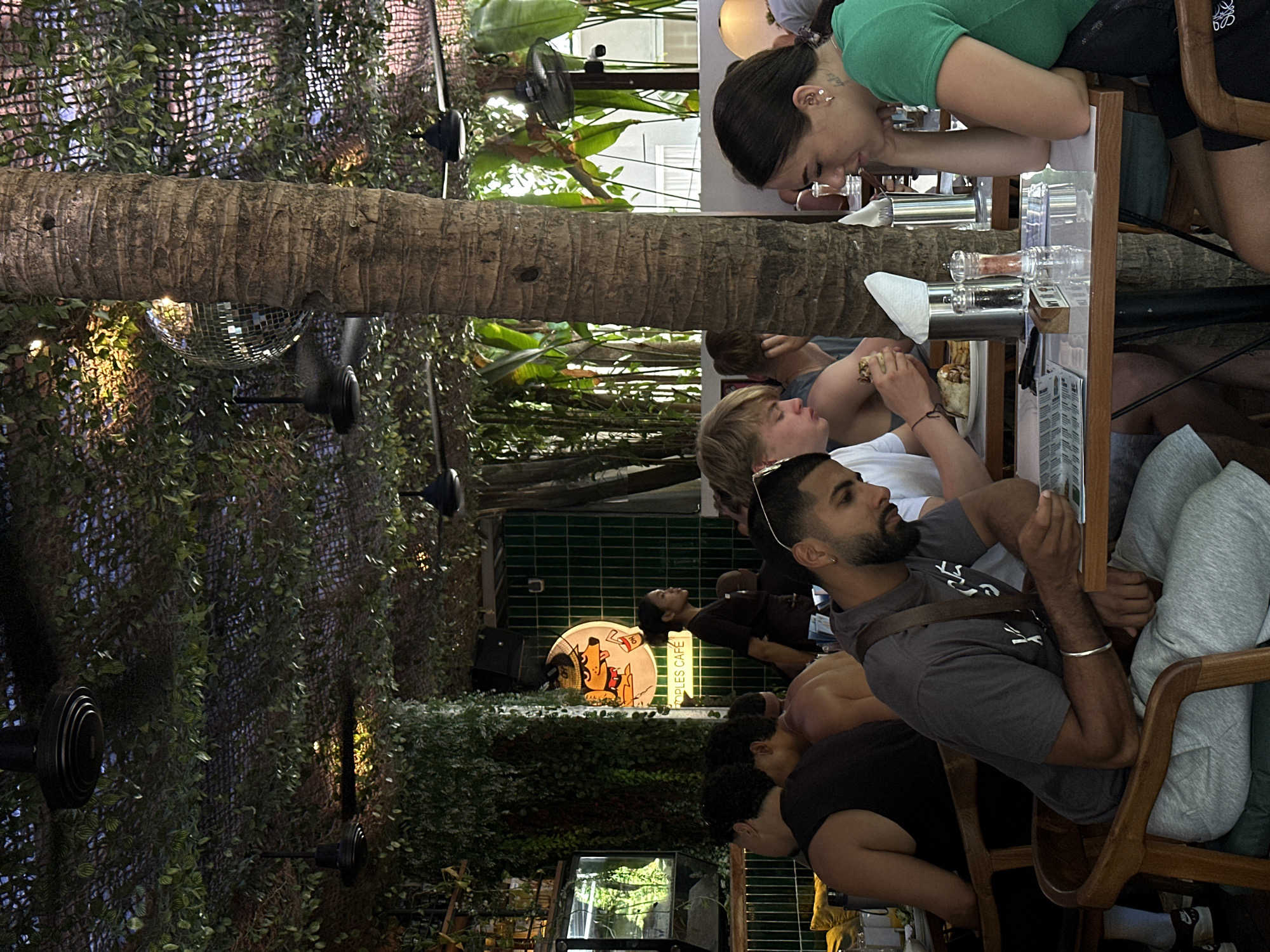 A group of young adults are seated at tables in an outdoor dining area, partially shaded by a canopy of greenery and a tree trunk. The setting appears to be "Peoples Cafe" in Mengwi, Indonesia, as indicated by a sign. On the left, a man in a gray t-shirt and gray sweatpants is looking down at a menu. Beside him, a person with blonde hair wearing a white t-shirt is eating a wrap. To the right, a woman with dark hair in a green shirt is looking down at her hands resting on the table. In the background, a disco ball hangs from the ceiling, and fans are visible, suggesting a tropical or warm climate. The overall atmosphere is casual and social, with people engaged in dining and conversation.