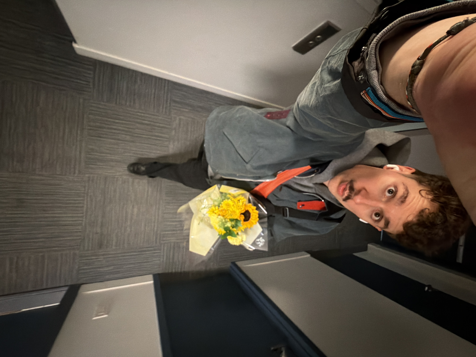 The image, taken from a high, overhead angle, shows a young man standing in an indoor hallway. The photograph is rotated, causing the man to appear sideways or inverted within the frame, but he is looking directly towards the camera's lens with wide, slightly surprised eyes. He has curly brown hair, a mustache, and a light beard. He is wearing a grey hooded sweatshirt underneath a blue-grey denim-like jacket, with an orange detail visible on his inner arm or collar. Dark trousers and black shoes complete his attire. White wireless earbuds are visible in his ears, and a multi-colored braided bracelet is on his right wrist.
He holds a vibrant bouquet of flowers, primarily yellow sunflowers and other smaller yellow blossoms, wrapped in light yellow paper and clear plastic. A dark backpack or bag is slung over his shoulder.
The setting is an indoor hallway, likely in a modern building such as an apartment complex or hotel. The floor is covered with dark grey carpet tiles arranged in a geometric pattern of alternating stripe directions. The walls are light grey or white, with a white baseboard running along them. A dark rectangular object, possibly a light switch or intercom panel with two buttons, is mounted on the wall behind him. Parts of dark doors are visible at the bottom and top edges of the frame. The lighting suggests an indoor artificial light source. No visible text is present in the image.