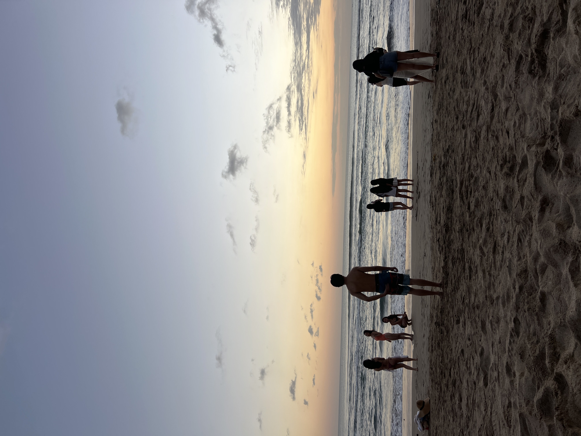 The image shows a group of people on Kuta Beach in Bali, Indonesia, at sunset. The sky is a gradient of blue, purple, and orange, with scattered clouds. The ocean is calm, with gentle waves rolling onto the shore. The sand is light brown and appears to be dry.
In the foreground, there are several people standing on the beach, facing the ocean. One man is in the center of the frame, wearing blue and black patterned swim trunks. To his left, there are three women. One is wearing a bikini, another is wearing a patterned swimsuit, and the third is crouched down. To the right of the man, there are two women standing closer to the water. They are both wearing dark tops and one is wearing denim shorts. Further to the left, a person is lying on the sand, and a hat and towel are placed next to them.
In the background, the ocean stretches to the horizon, meeting the colorful sunset sky. The overall atmosphere is serene and peaceful, capturing a moment of leisure on a popular Indonesian beach.