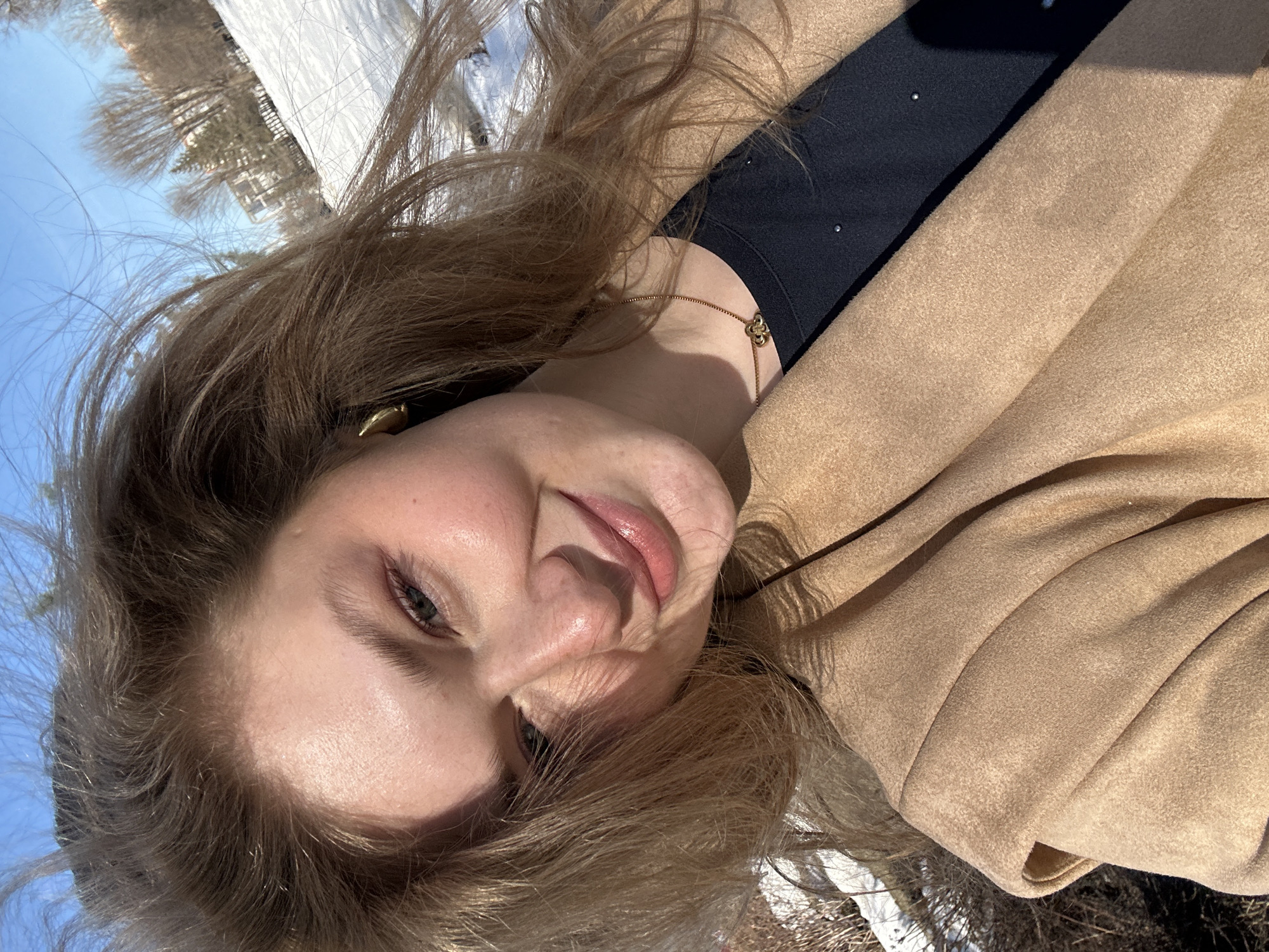 A medium close-up of a woman with blonde hair, wearing a beige coat and a black top, possibly taken in Kazan, Russia (based on a location request that is separate from the image itself). The sky appears blue. The woman is looking at the camera. Part of the background contains a blurred view of bare trees and possibly snow. The image orientation is at an angle, with the top of the frame showing a blue sky and blurred trees and a white surface.