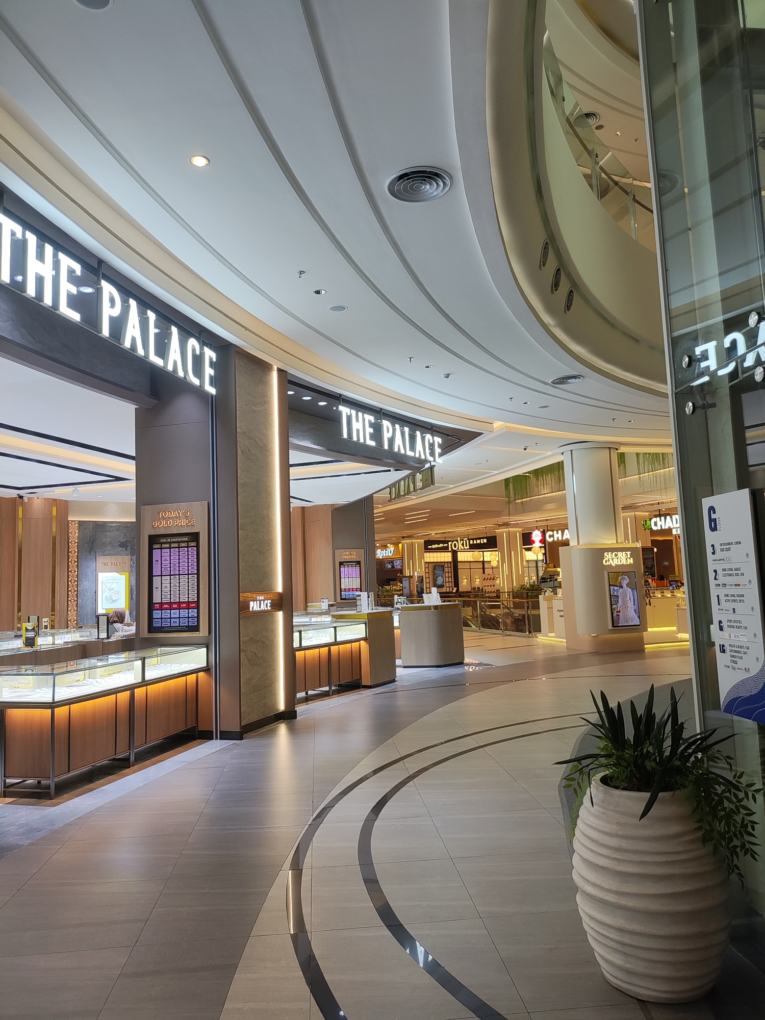 The image shows the interior of a modern shopping mall. The scene is well-lit with a combination of overhead lighting and accent lighting from the shops. In the foreground, a large, textured white planter holds several green plants, casting shadows on the tiled floor. The floor itself is made of large, light-colored tiles, with curved darker gray lines creating a pathway.
Dominating the left side of the image is a storefront with a large illuminated sign that reads "THE PALACE." Below the sign, a display features jewelry in glass cases with warm wooden cabinets. A digital screen showing "TODAY'S GOLD PRICE" is also visible. To the right of "THE PALACE" storefront, another similar storefront with the same name is partially visible, also featuring illuminated signage and glass display cases.
In the background, the mall's architecture is characterized by curved ceilings and structural elements. Various other shopfronts are visible, including one with a sign for "ROKU RAMEN" and another for "CHAD." A large pillar with the "SECRET GARDEN" logo and an image of a dress is also present. A directory sign with the letter "G" and various floor listings is visible on the far right. The overall environment suggests a clean, upscale retail setting. No people are clearly identifiable in the scene. The location is identified as Lambangjaya, Indonesia.