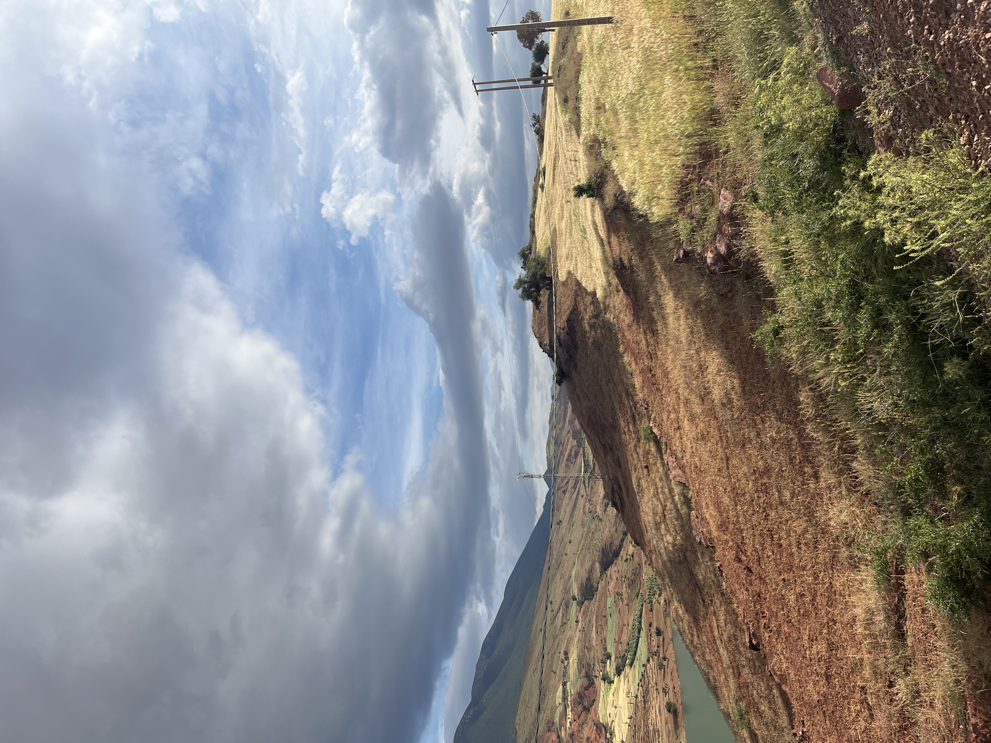 The image shows a landscape in Ouaouizert, Morocco. The foreground features a steep slope covered in dry, brown vegetation and some patches of green foliage. At the top of the slope is a dirt path, then a field of dry grass with utility poles and power lines. In the background, a mountain range is partially obscured by cloudy skies. Below the mountains, there are agricultural fields with a network of irrigation channels. The sky is filled with a mix of dark, stormy clouds and patches of blue sky.