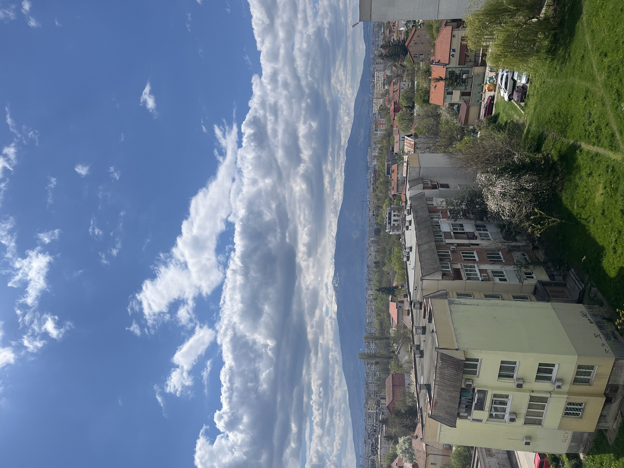 The image presents a panoramic view of a city, likely Sofia, Bulgaria, with a prominent mountain range in the background, all set under a bright, partly cloudy sky. The photograph is oriented vertically, but depicts a horizontal landscape.
The upper portion of the scene (left side of the rotated image) is dominated by a clear blue sky interspersed with numerous white clouds. These clouds, appearing as cumulus and stratocumulus formations, stretch in long, somewhat parallel streaks across the sky, suggesting good weather conditions.
In the mid-ground, a large mountain range, consistent with Vitosha Mountain, extends across the horizon. Patches of white, indicative of snow, are visible on some of the higher peaks. Below the mountains, the urban sprawl unfolds, characterized by a dense collection of residential buildings. These include multi-story apartment blocks and houses, many featuring traditional red-tiled roofs, alongside structures with flat roofs, showcasing varied architectural styles.
The foreground (right side of the rotated image) focuses on closer residential areas and green spaces. A distinctive, light yellow-green multi-story building stands out in the lower right, equipped with multiple windows and exterior air conditioning units. Laundry is visible drying on a line attached to this building. Adjacent and further right are vibrant green grassy areas, some traversed by visible dirt paths. Several trees are scattered throughout the scene; one near the yellow-green building is in bloom with white flowers, and a large weeping willow is prominent in the far right. A few cars, including white and dark red models, are parked on a grassy patch in the mid-ground.
The time of day appears to be late morning or early afternoon, given the bright, even illumination and distinct shadows. On the yellow-green building, Cyrillic text is visible. The letters "ЧСK" are clearly legible on the lower facade. Higher up, on a visible side, fragmented text and symbols include "УКР," suggesting graffiti or a message, possibly related to "Україна" (Ukraine). The overall scene captures a calm, everyday urban environment nestled against a natural mountainous backdrop.