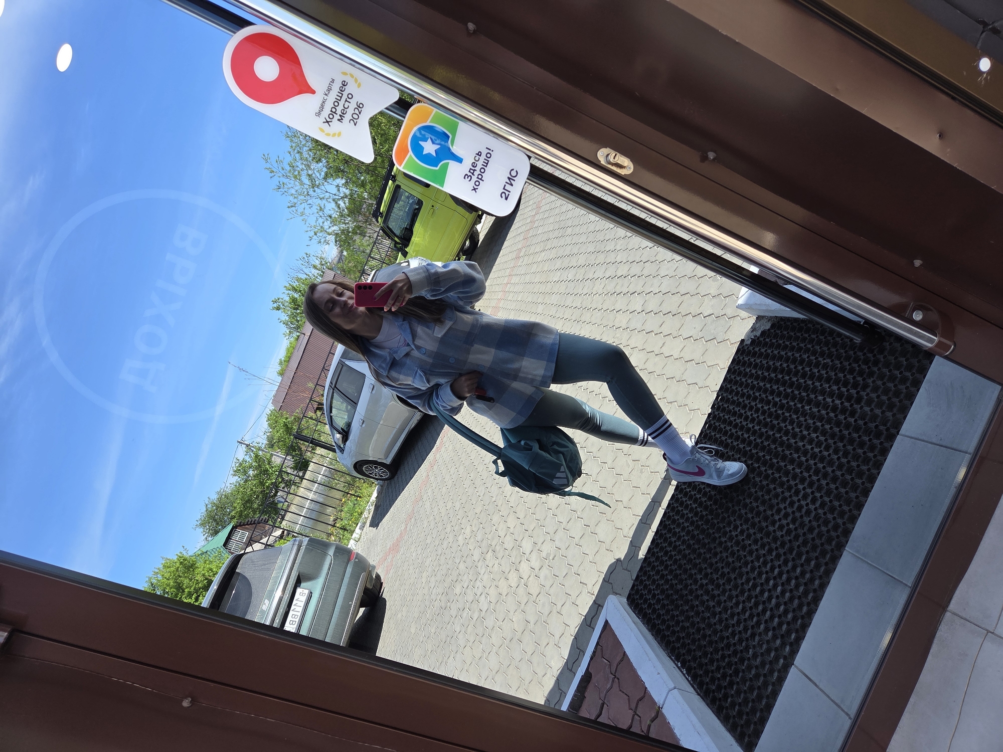 A young woman is taking a selfie in a doorway, reflected in the glass. She is wearing a plaid shirt, leggings, and sneakers, and carrying a backpack. The location is Birobidzhan, Russia, indicated by the two stickers on the door that say "Good place" in Russian and show the 2GIS logo. Outside, there is a paved area with parked cars and trees. The sky is blue and sunny.