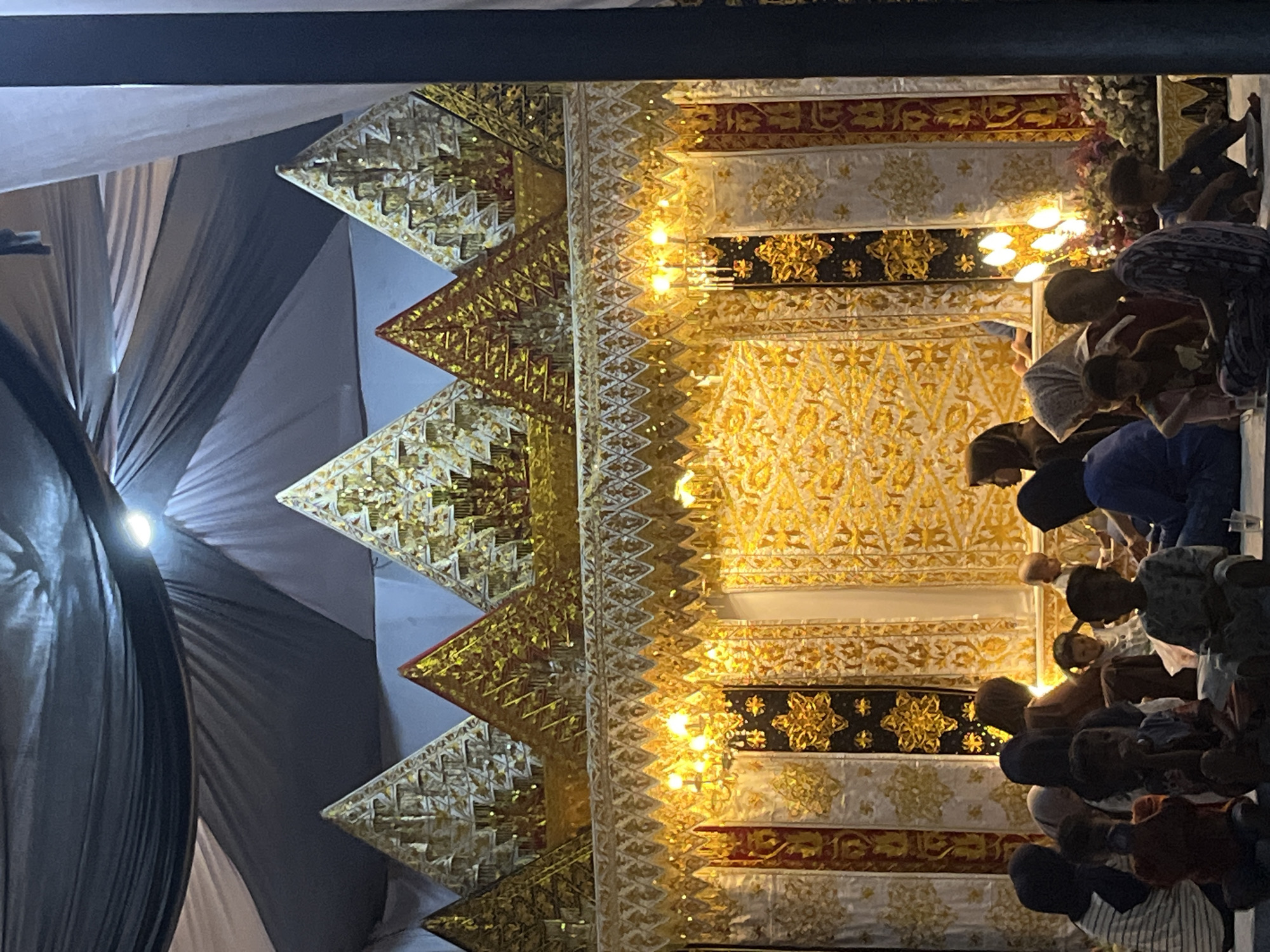 This image captures an evening gathering within an ornate, likely temporary, setting in Lhokseumawe, Indonesia. The scene is dominated by a grand, elaborately decorated structure on the left side of the frame (when oriented vertically). This structure features multiple layers of intricate patterns in gold, white, black, and touches of red, with distinct, stepped, triangular golden elements forming its left edge. Small, warm-toned lights are embedded throughout this structure, illuminating its detailed motifs and casting a soft glow. This opulent backdrop suggests a ceremonial or religious function, possibly resembling a mihrab or a decorated stage.
To the right of this structure, a group of people, including many children and some adults, are gathered. They are seated or kneeling on the floor, mostly with their heads bowed or looking forward in a reverent posture. Their attire appears modest and suitable for a formal or religious event.
In the background on the far left, large swaths of dark gray fabric are draped, illuminated by a bright light source from behind or within, suggesting a temporary enclosure like a tent or a specially decorated hall. The artificial lighting indicates that the event is taking place at night or in the late evening. The overall atmosphere appears solemn and focused. No discernible text is visible in the image.