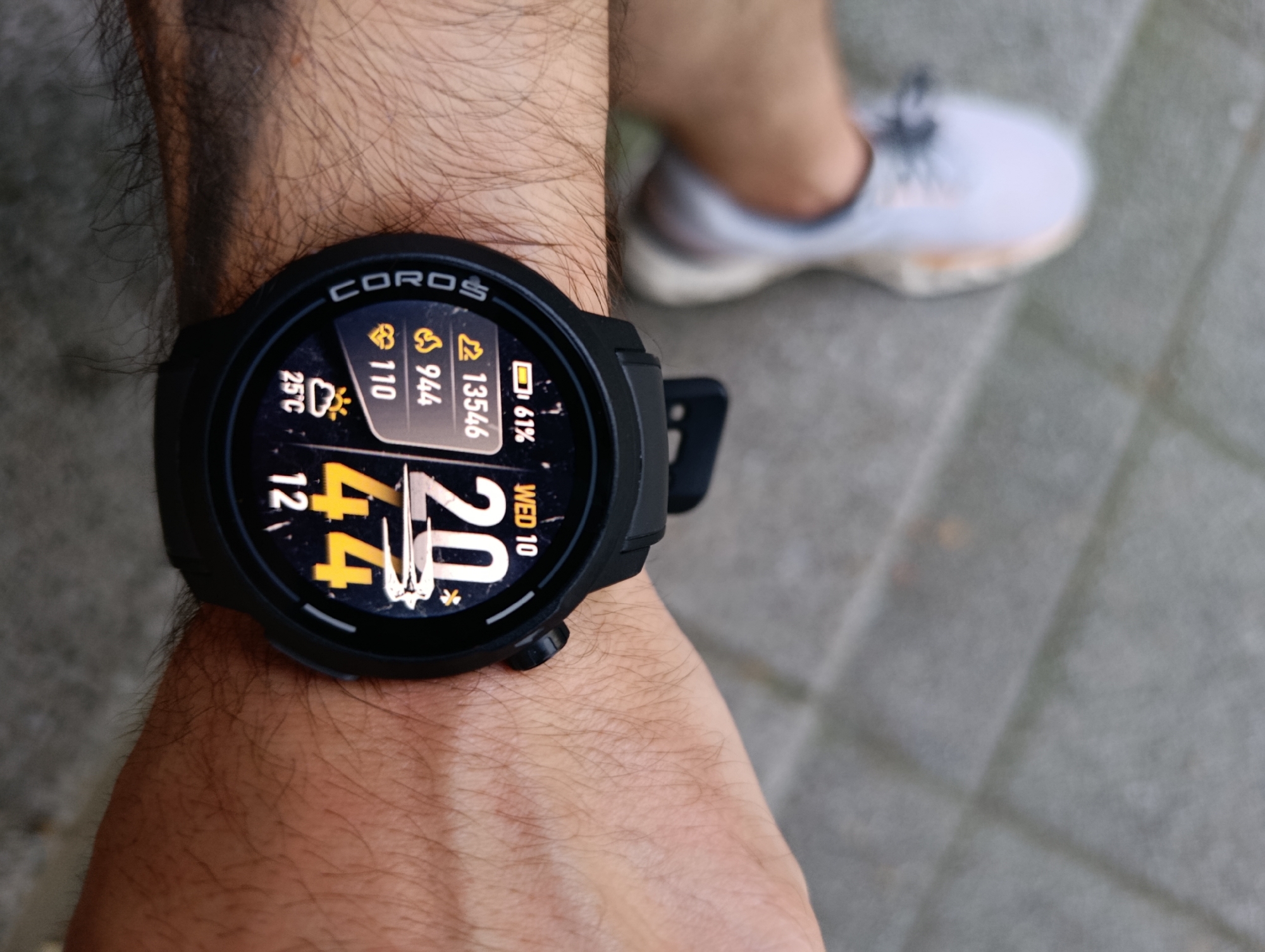A close-up, eye-level shot shows a person's wrist with a black COROS smartwatch. The watch face displays weather information, including a cloud and sun icon with "25°C" below it. To the right, there are icons for steps (a shoe) and a battery level, showing "110," "944," "13546," and "61%." The time is displayed as "4:42" and "20," with "WED 10" below the latter. The background is blurred and shows a paved surface, likely a sidewalk, and a person's foot wearing a light-colored athletic shoe. The scene is outdoors, with the lighting suggesting daylight.