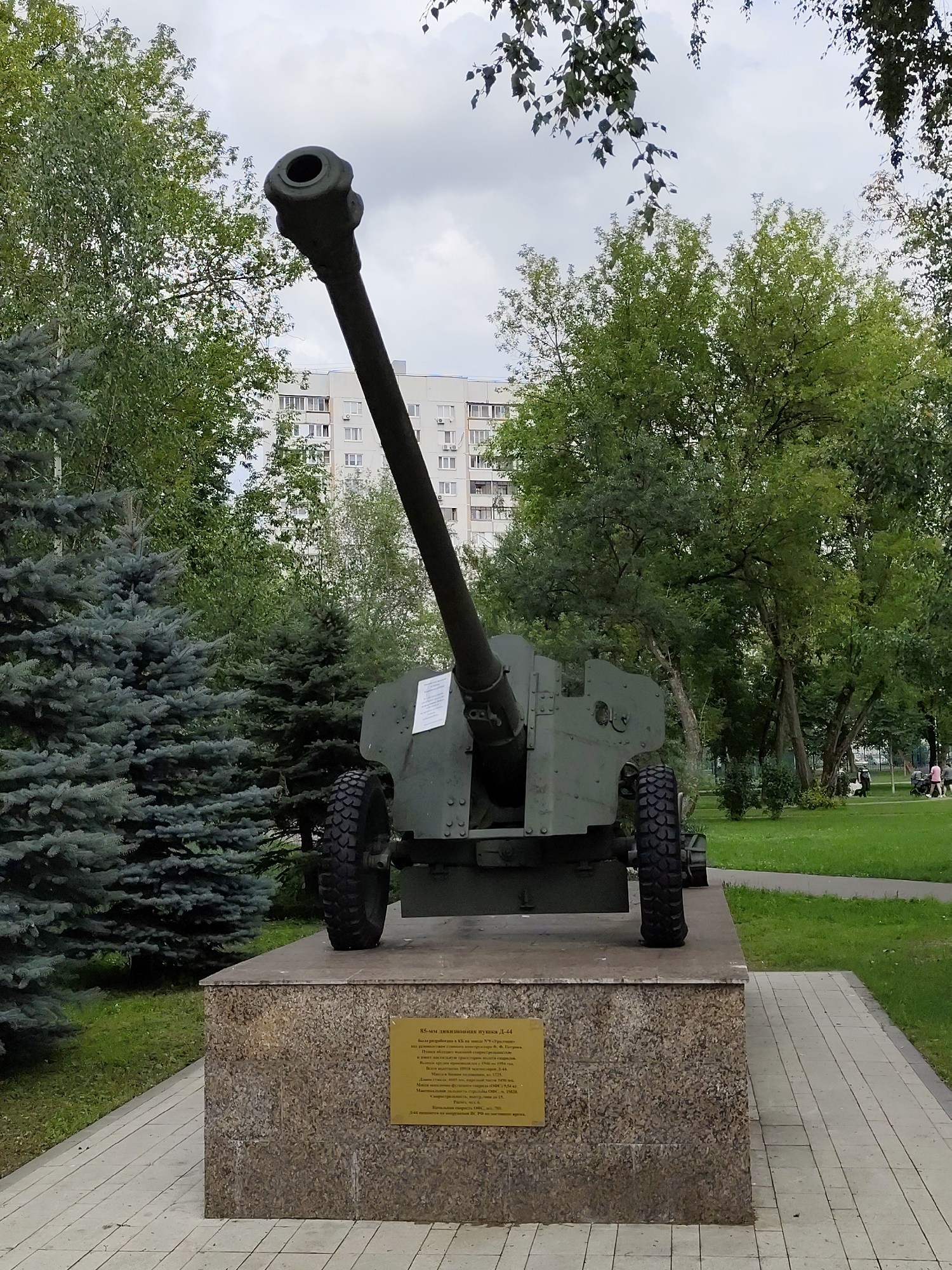 An anti-tank gun is displayed on a stone pedestal in a park. The gun is painted olive drab and has a long barrel. The pedestal is covered in a granite-like material. Behind the gun, there are trees and a tall, white apartment building. In the background, there are a few people walking in the park. The sky is overcast. The anti-tank gun is a D-44, a Soviet 85 mm divisional gun. This gun is located in Moscow, Russia, specifically in Izmailovsky Park.