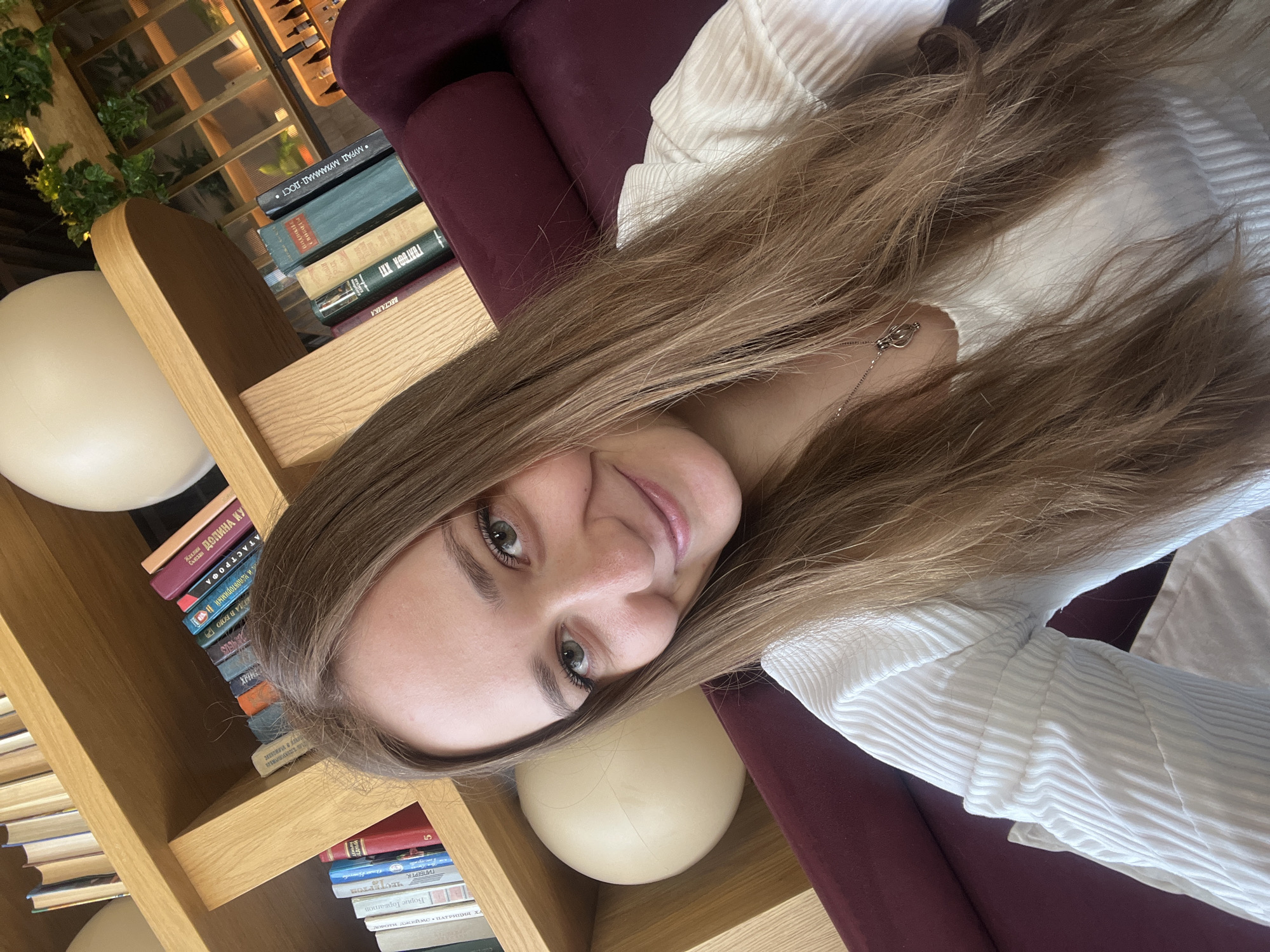 A selfie taken indoors features a woman with long brown hair, light skin, and green eyes. She is wearing a white ribbed shirt. She is sitting on a burgundy upholstered chair or couch. To her left is a wooden bookcase filled with books of various sizes and colors, with two round, cream-colored lamp-like objects on the shelves. Some of the titles on the books are visible and written in what appears to be Russian. The background shows glimpses of what looks like a window or a decorative structure, possibly with plants. The location is Verkhnyaya Pyshma, Russia.