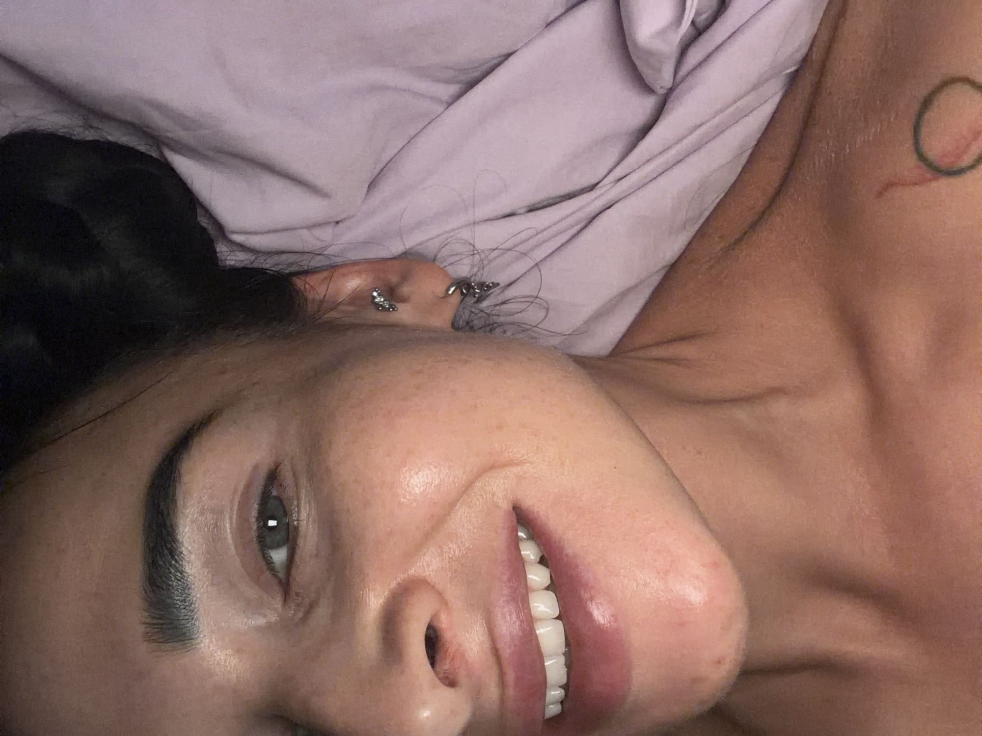 The image presents a close-up portrait of a person, likely female, lying horizontally, possibly in bed. The subject is smiling gently and looking directly at the camera with an engaging, relaxed expression. Her dark hair is visible on the left side of the frame, with some loose strands near her ear.
Her skin is fair, with subtle freckles visible on her cheek. She has prominent, dark eyebrows and light-colored eyes. A gentle smile reveals her healthy, white teeth. Multiple metallic piercings adorn her ear, including small studs and more intricate hoops. On her right shoulder or upper chest area, a faint tattoo resembling an incomplete circle or loop with a reddish element is visible.
A rumpled light lavender or pale purple sheet covers part of her body and forms the background, suggesting an indoor setting, likely a bedroom. The lighting appears soft, highlighting her features and the texture of the sheet. The overall mood is intimate and casual. No visible text is present in the image. The scene is set in Saint Petersburg, Russia.