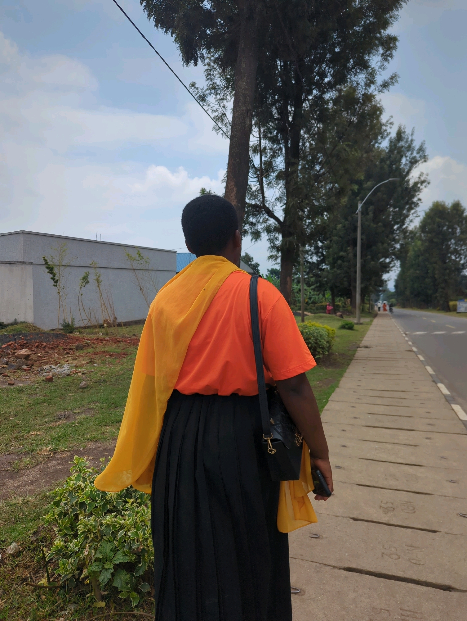 A person wearing an orange t-shirt, a yellow shawl, and a black pleated skirt walks along a concrete sidewalk next to a road. The person is carrying a black purse over their left shoulder and holding a dark-colored mobile phone in their right hand. The sidewalk is lined with trees on the left and a paved road on the right. In the background, there is a building with a light-colored facade, some vegetation, and more trees. The sky is partly cloudy. The location is identified as Musanze District, Rwanda.