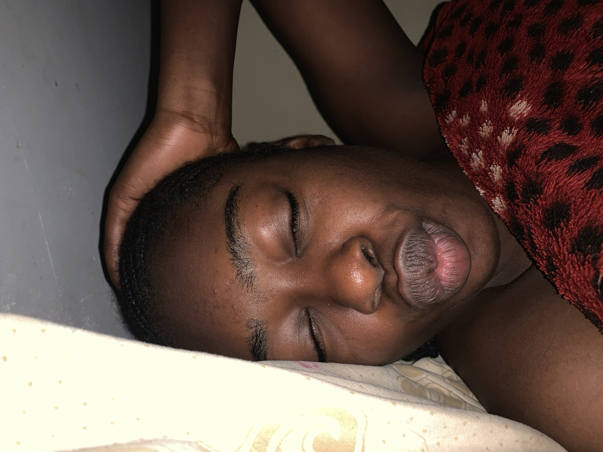 The scene depicts an indoor, low-light environment in Rafin Par, Nigeria. A dark-skinned individual lies on their right side, eyes closed, indicating sleep. Their head rests on a cream-colored pillow featuring a subtle light-toned pattern. The individual has short, dark, closely-cropped hair. Their facial features include a broad nose and full lips, which are slightly parted, revealing a lighter reddish hue on their inner surface. A red blanket with distinct irregular dark spots, resembling a leopard print, covers a portion of their upper body. The blanket exhibits a plush, textured surface with visible lighter-colored fibers. Above and behind the sleeping individual's head, a dark-skinned forearm and hand are visible, with the hand gently resting near the individual's scalp. The background consists of a plain, light gray wall. The overall lighting is dim, casting soft shadows.