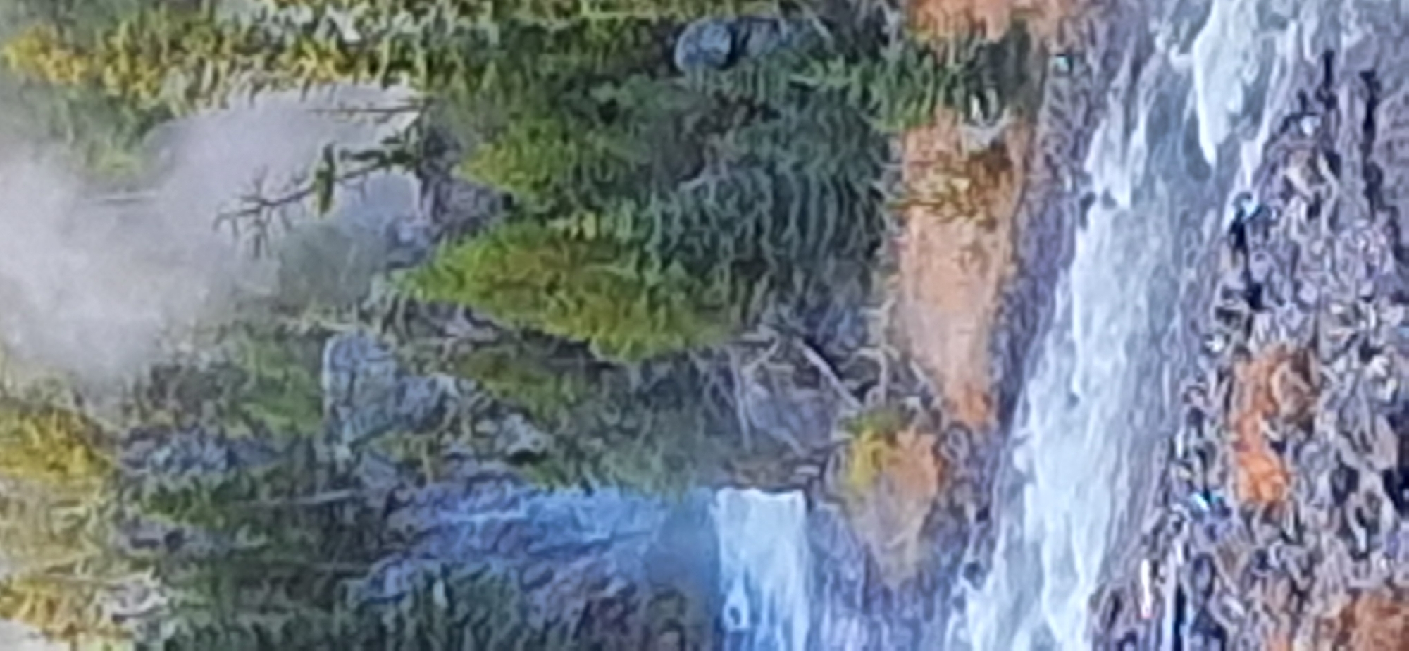 The image depicts a natural landscape featuring a waterfall cascading into a river. Dense evergreen trees line the rocky banks, with mist rising in the background. The foreground shows a rocky riverbed. There are no people or urban elements visible, suggesting a wilderness setting. This scene is not located in Bila Tserkva, Ukraine, as there are no identifiable landmarks or features corresponding to that city.