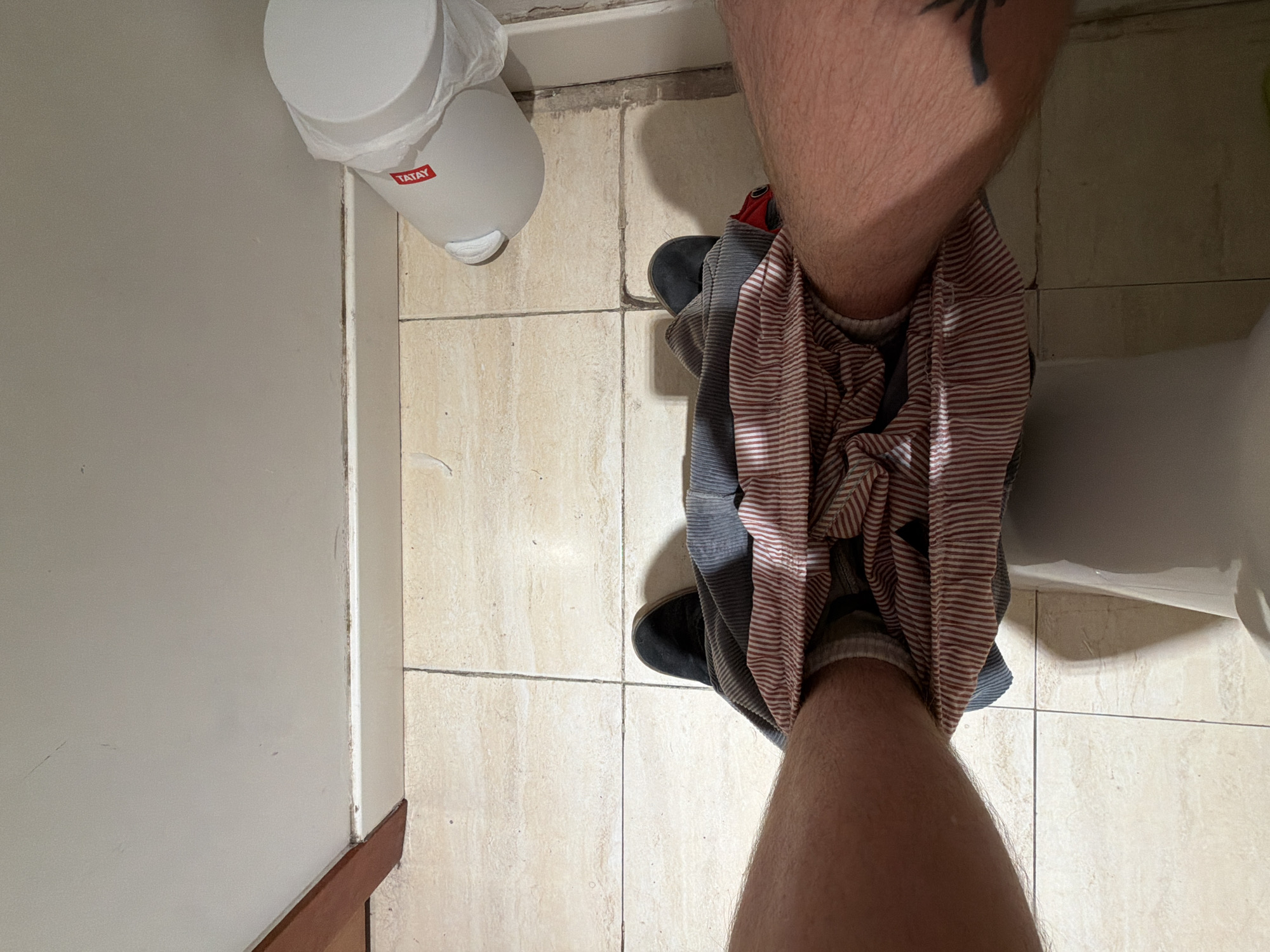 A first-person, overhead shot depicts a person with their pants pulled down around their knees, standing in a bathroom. The person's legs are visible, with a tattoo on the upper left leg. The person is wearing grey socks and black slip-on shoes. The pants are striped grey and red. To the left is a white wall with a brown baseboard. Above the person is a white trash can with the word "Tatay" printed on it. A white toilet is also visible to the right. The floor is covered with beige square tiles. There is no clear indication of the location being Barcelona, Spain.