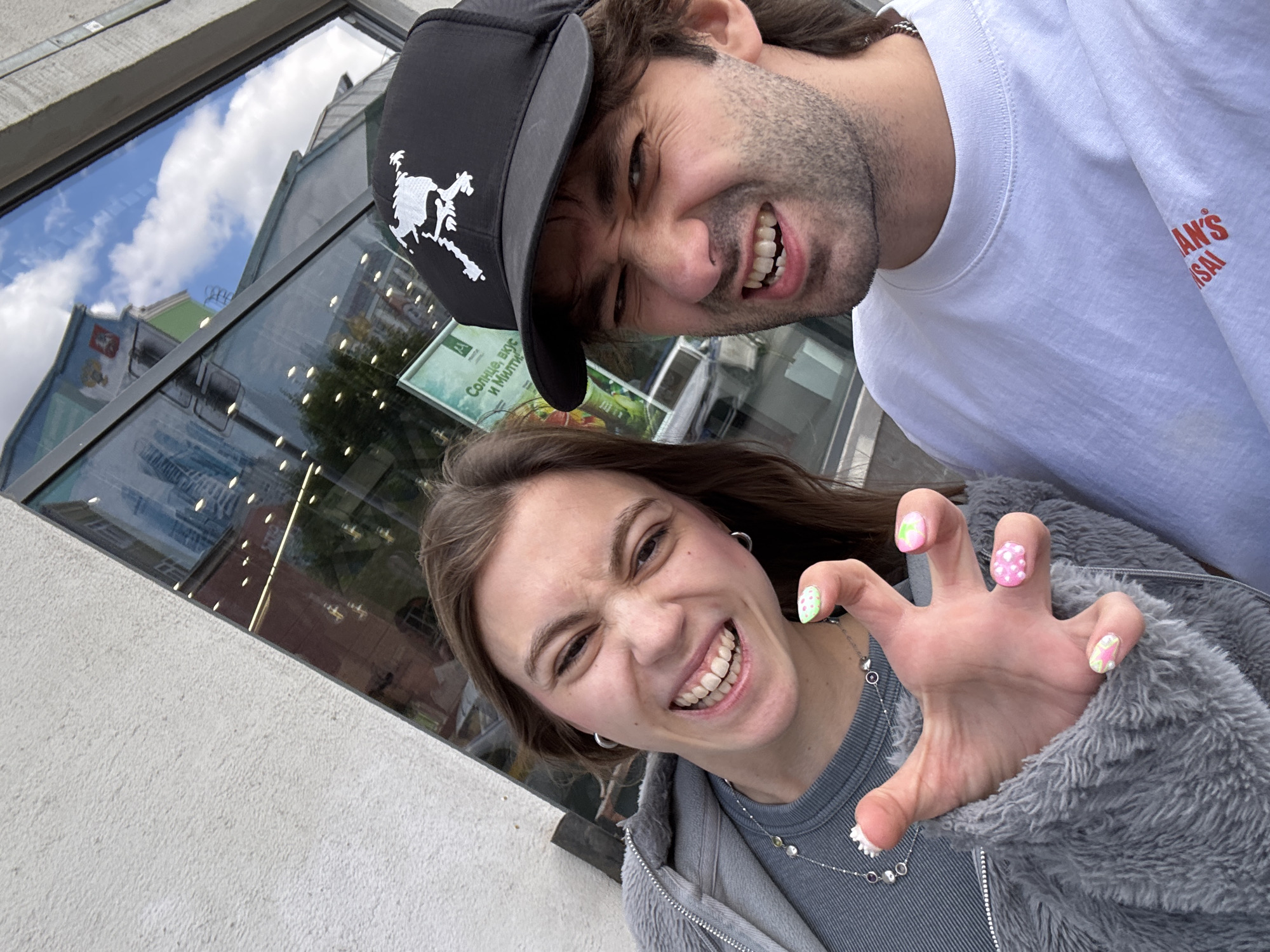 This is a selfie taken by two people, a man and a woman, in an outdoor urban setting. The man is in the foreground, wearing a black baseball cap with a white graphic and a white t-shirt. He is making a distorted facial expression, with his eyes squinted and his mouth open in a wide grin, showing his teeth. The woman is positioned slightly behind and to the side of the man, also looking at the camera. She has brown hair and is wearing a grey zip-up hoodie. She is also smiling broadly and has her hand raised in front of her face, with her fingers splayed in a claw-like gesture. Her nails are painted with colorful polka dots and other patterns.
The background appears to be a building with large glass windows. Reflections of the sky, clouds, and other buildings are visible in the glass. There is a sign visible through the glass with some text that appears to be in Russian. The overall mood of the photo is playful and energetic, indicated by the exaggerated facial expressions and the woman's pose. The lighting suggests it is daytime, and the weather appears to be clear, as indicated by the visible sky and clouds. The presence of a building with windows and reflections suggests an urban environment, and the context provided states the location is Moscow, Russia.