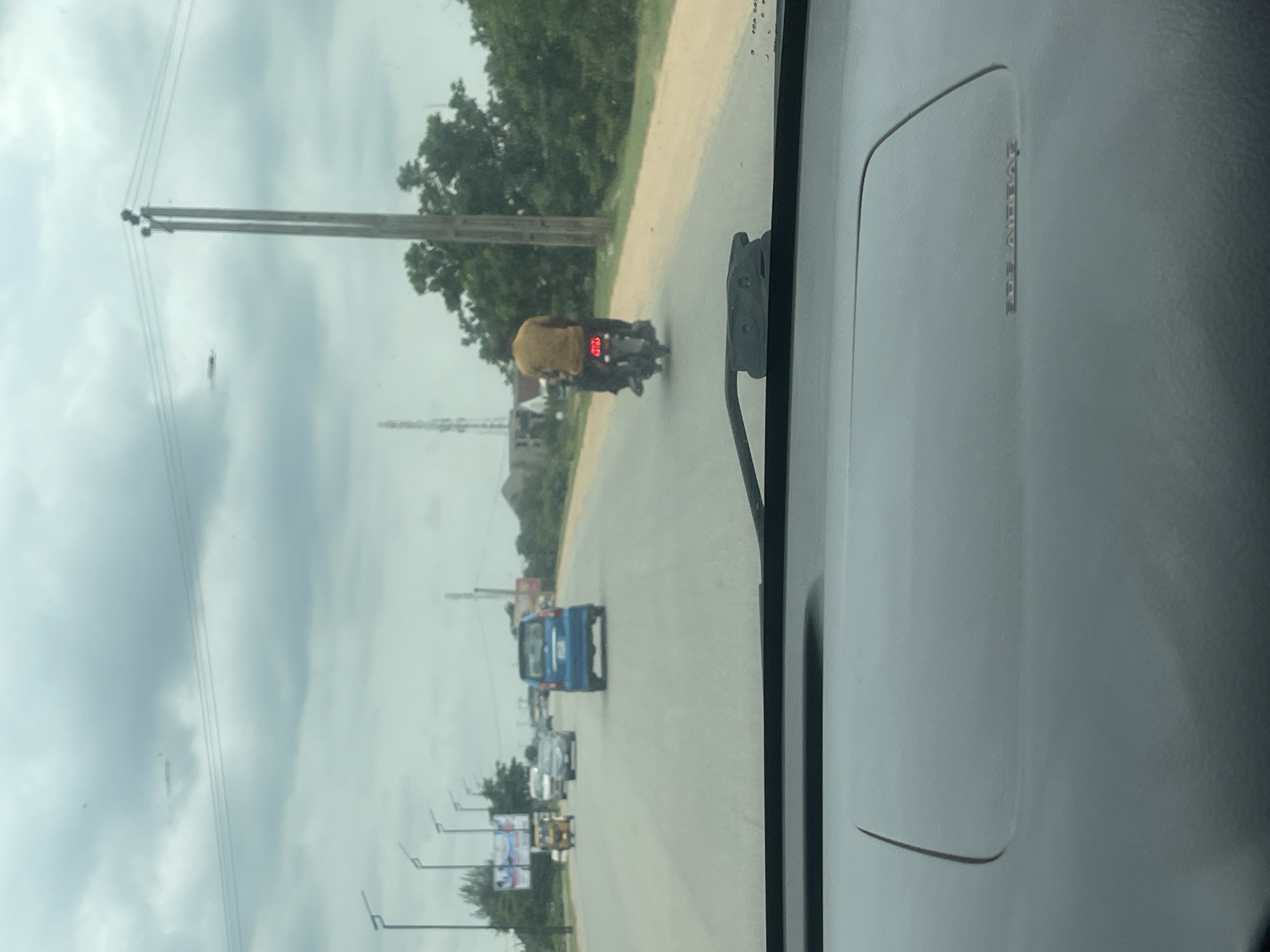 The image captures a view from inside a vehicle looking out onto a road in Galadimawa, Nigeria. The sky is overcast with heavy clouds. On the road, a blue car is traveling away from the camera, and a motorcycle with two occupants is also moving forward. The driver of the motorcycle is wearing a yellow shirt, and the passenger is wearing dark clothing. The road is paved, with a dirt shoulder on the right side. Trees and vegetation line the side of the road, and utility poles are visible in the distance. The foreground of the image shows the dashboard of the vehicle, including the "AIRBAG" warning.