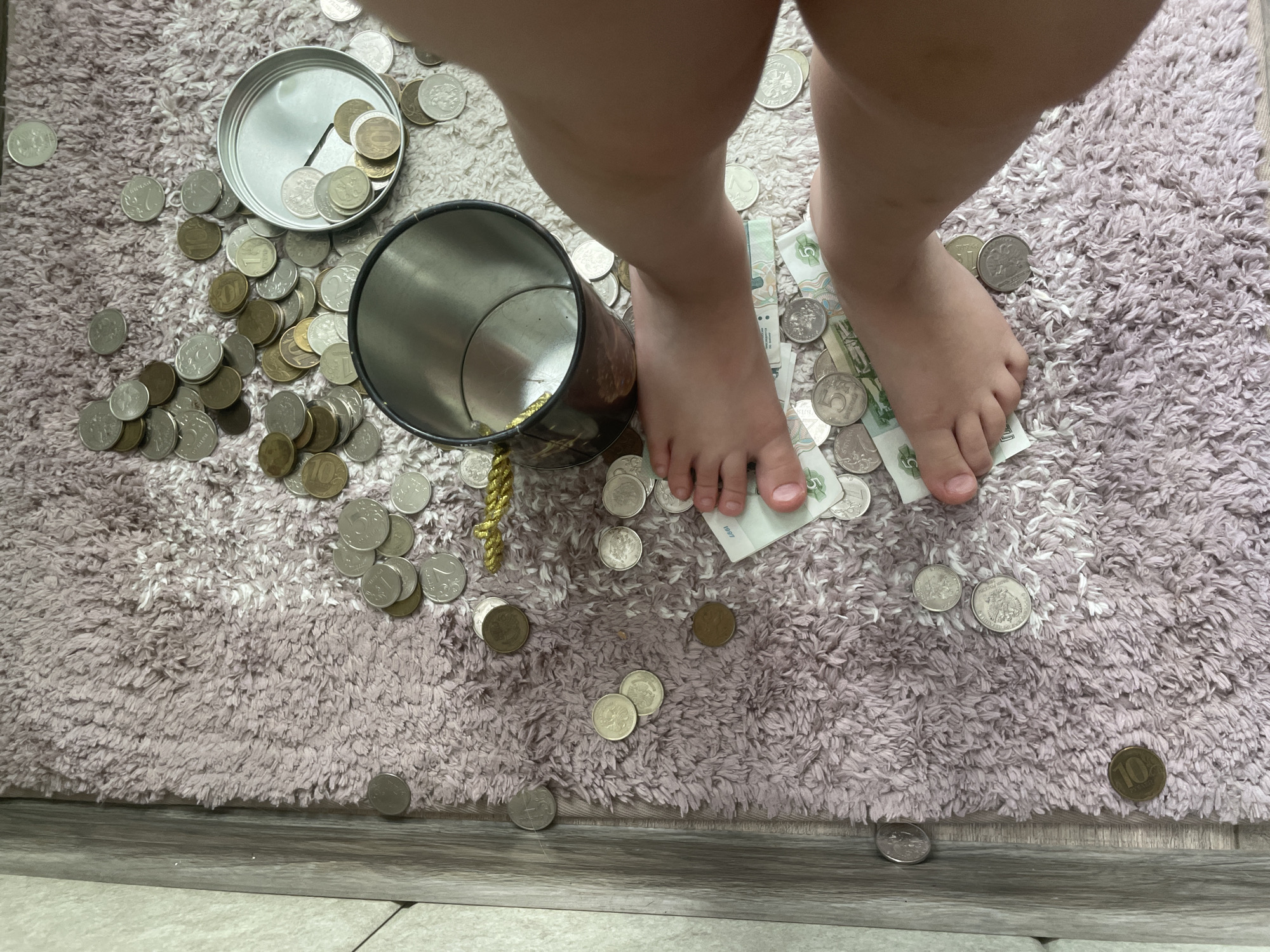 This is a top-down, eye-level, full shot of a child's bare feet and legs resting on a plush, light pink rug. The child is surrounded by a large scattering of Russian coins and several Russian banknotes. The coins are of various denominations and are spread haphazardly across the rug, some clustered together, others isolated. A few coins are inside two open metallic containers, one of which is partially visible at the top right of the frame. The banknotes are mostly obscured by the coins and the child's feet, but some have denominations of "5" visible. The child's feet are positioned centrally, with toes pointed upwards. The environment suggests an indoor setting, possibly a child's room. The image does not provide any information about the city of Vladivostok, Russia.