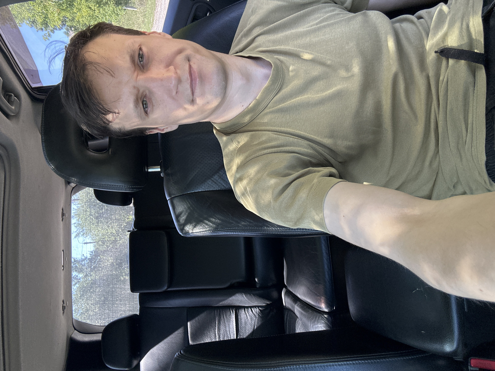A man is sitting in the driver's seat of a car, looking at the camera. He has short, dark brown hair and light green eyes. He is wearing a light olive green t-shirt. The interior of the car is black leather, and the back seats are visible behind him. The car appears to be parked on a dirt road, with trees and a blue sky visible through the windows. The image is taken from a first-person perspective, as if the man is taking a selfie. The man is in the city of Makiv, Ukraine.