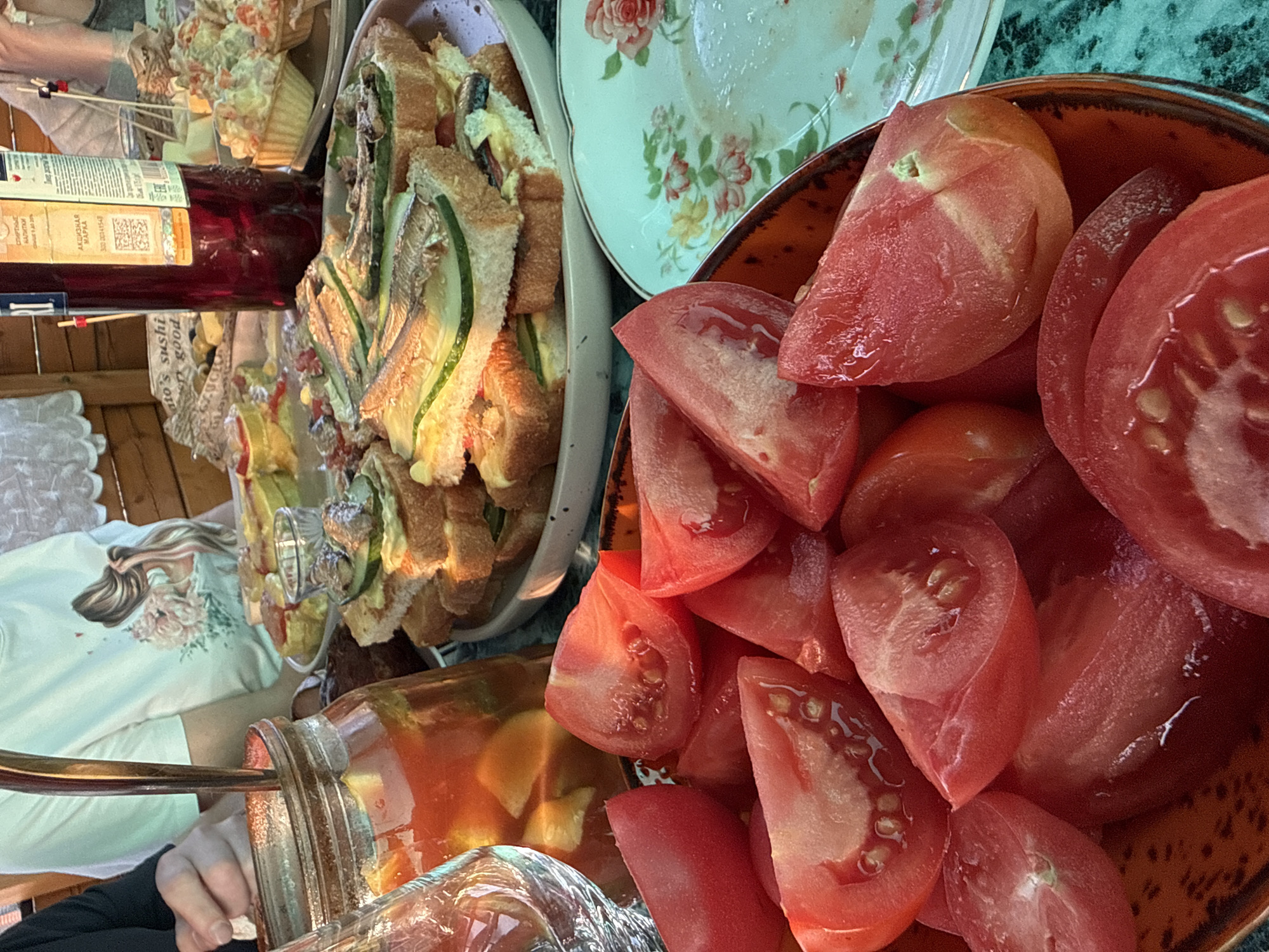 A close-up shot captures a table laden with food. In the foreground, a rustic orange bowl overflows with bright red, juicy tomato wedges, glistening with moisture. To the right, a delicate floral-patterned plate, likely ceramic, appears partially used, with remnants of food. Further back, a platter holds an arrangement of small open-faced sandwiches, topped with sliced cucumbers and small fish, possibly anchovies. Behind the sandwiches, a tall, dark red bottle, possibly wine or aperitif, stands upright. A glass jar filled with a vibrant red liquid, containing fruit slices and ice, sits to the left. A partially visible clear glass is also present. The background suggests an outdoor setting with wooden elements and a white tablecloth. A person's arm and hand are visible on the left side, reaching towards the jar. The overall scene depicts a casual outdoor meal or gathering. The location is not identifiable from the image.