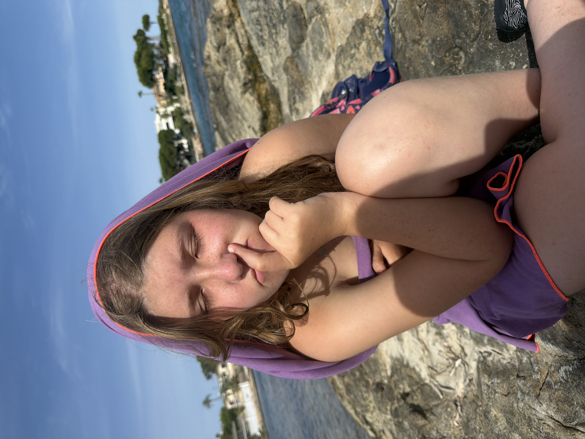The image shows a young person lying on a rocky surface by the sea, with a scenic view of a coastline in the background. The person is wearing a purple hooded towel, with orange trim, draped over their head and shoulders, and a purple swimsuit. Their eyes are closed, and their finger is touching their nose, suggesting a relaxed or contemplative mood. The scene is bathed in bright sunlight, indicating it is likely daytime. The background features clear blue sky and water, with buildings and trees visible along the distant shore, characteristic of a Mediterranean coastal town. The rough texture of the rocks and the sparkling water suggest a natural, outdoor setting, possibly a beach or a rocky cove. The overall impression is one of a peaceful moment during a day at the seaside in a sunny location.