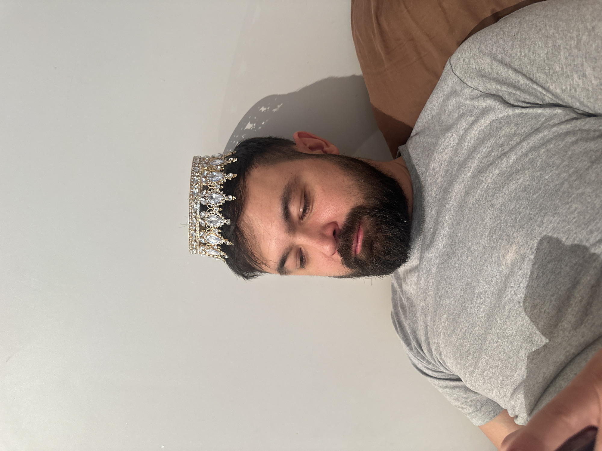 An adult man with a beard and dark hair is depicted sleeping or resting, lying horizontally against a plain, light-colored wall. His eyes are closed, and his expression appears peaceful. He is wearing a grey short-sleeved t-shirt.
A prominent gold-toned crown, richly embellished with numerous clear, sparkling jewels, rests on his head. Part of a brown object, likely a pillow or blanket, is visible behind his head and right shoulder, suggesting he is lying on a soft surface. The background is simple and uncluttered, consisting solely of the light wall. Shadows cast on the wall behind his head and crown suggest an indoor setting with an overhead light source. The overall scene conveys a moment of quiet repose, possibly with a touch of humor or irony due to the crown. No specific time of day or outdoor weather can be discerned. The image context indicates this media file is from Moscow, Russia. There is no visible text.