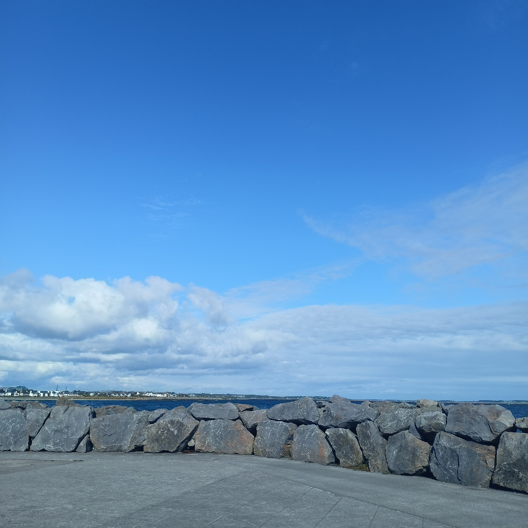 The image depicts a clear blue sky with scattered white clouds. In the foreground, a paved surface transitions to a substantial breakwater constructed from large, grey, irregularly shaped rocks. Beyond the breakwater, the dark blue ocean stretches to the horizon, where a low-lying town with white buildings and some cranes is visible, identifiable as Salthill, Galway, Ireland. The overall scene is one of a coastal landscape under a bright sky.