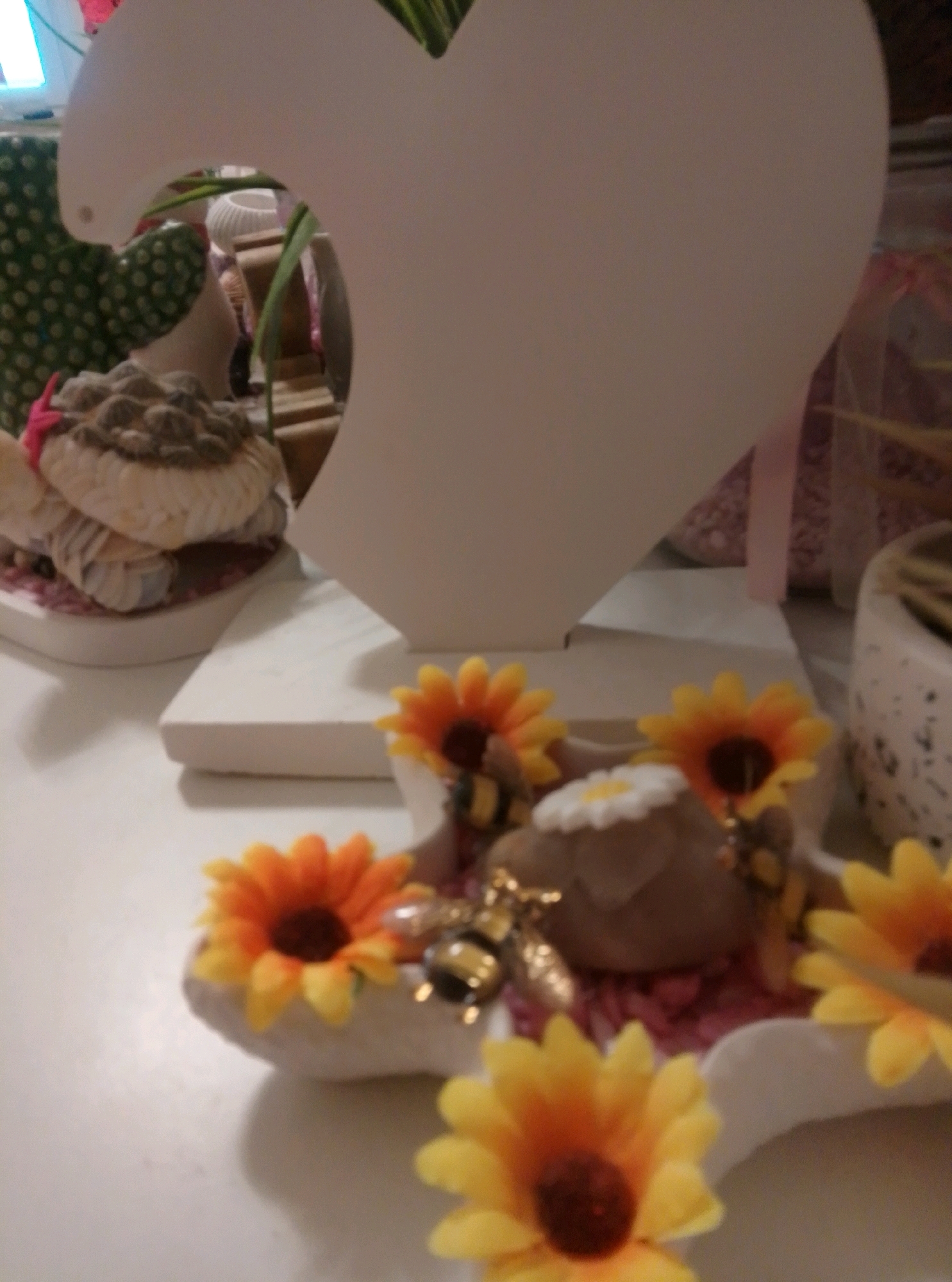 A shallow depth of field photograph shows a display of decorative items, likely for sale in a shop in Wedel, Germany. In the foreground, on a white surface, are several artificial sunflowers with orange and yellow petals and dark centers. Some of these sunflowers are nestled in small, decorative white containers. A few small, metallic bee figurines are interspersed among the flowers. Behind the sunflowers, a larger, white, heart-shaped object stands upright. To the left of the heart, a stack of what appear to be decorative items wrapped in fabric, possibly a gift or part of a display, is visible. The background is blurred, showing hints of other objects and colors, including what looks like a white ceramic pot and some pink material. The lighting is soft, and the overall impression is of a retail display.