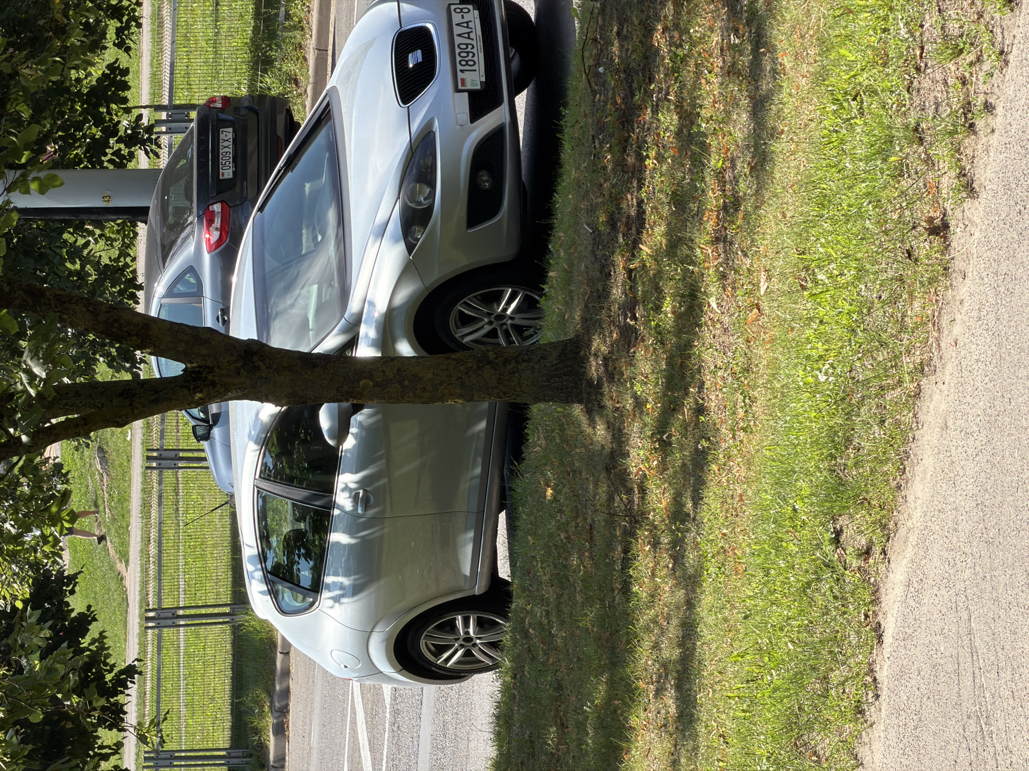 The image shows two cars parked near a grassy area in Minsk, Belarus. The foreground car is a silver hatchback, possibly a Seat Leon, with a license plate that reads "1899 AA-8". A tree partially obscures the view of the car. In the background, a dark-colored sedan is parked behind the silver hatchback, with its license plate reading "0509 XX-7". A metal fence runs along the edge of the grassy area, with a road visible beyond it. The environment suggests a sunny day, with dappled sunlight falling on the grass and cars. A person's legs are visible in the distance, walking on a path.