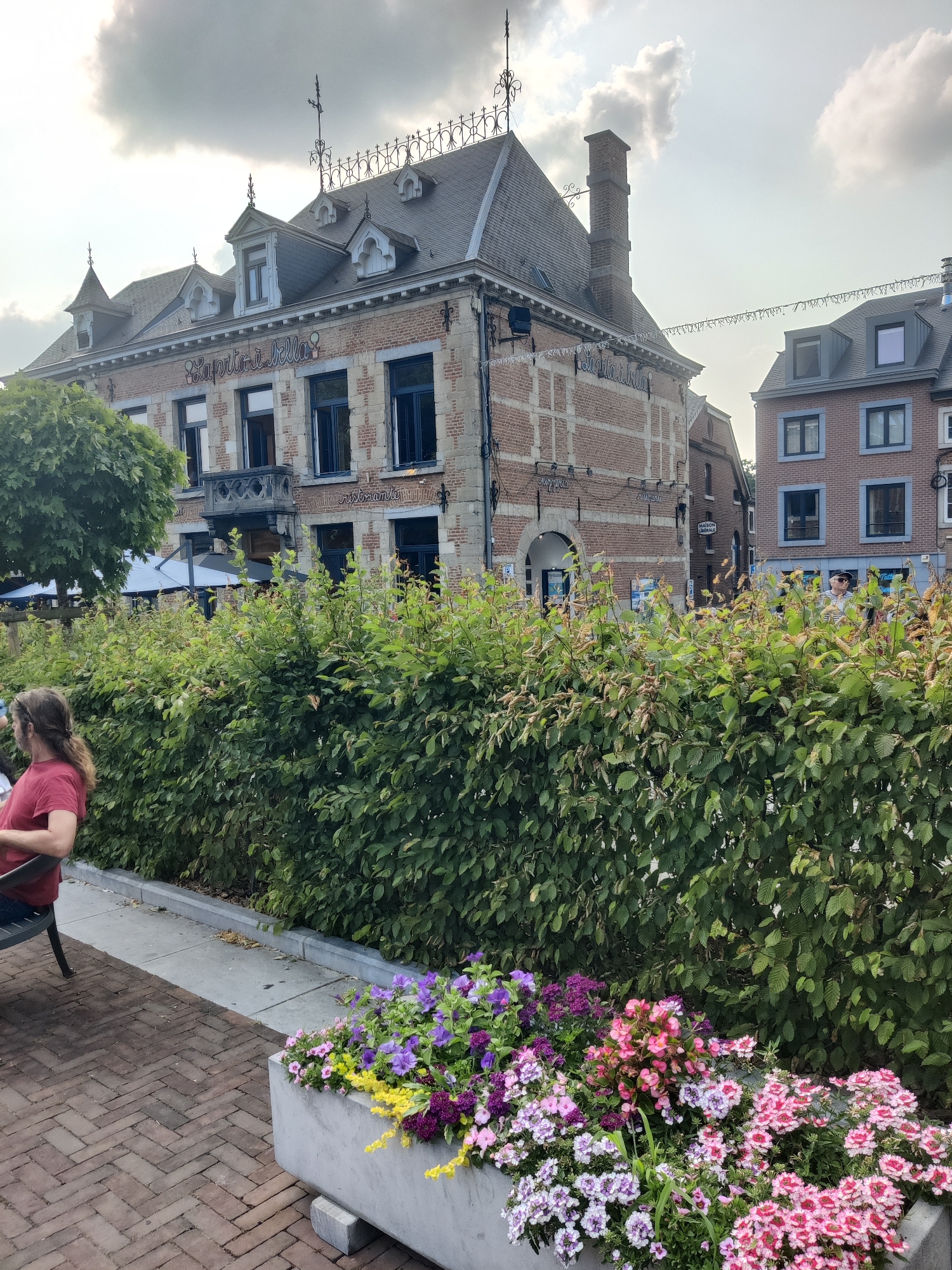 The image depicts a street scene in Marche-en-Famenne, Belgium. In the foreground, a concrete planter overflows with a vibrant arrangement of pink, purple, and yellow flowers. Behind the planter, a thick, well-maintained green hedge runs horizontally across the frame. To the left, a person with long brown hair, wearing a red shirt, sits on a dark bench, facing away from the camera. The ground is paved with red brick in a herringbone pattern.
Dominating the mid-ground is a multi-story building with a red brick facade and stone accents. A sign above the first floor windows reads "La Partigiana". The building has a pitched roof with dormer windows and ornate detailing along the roofline. A balcony with decorative ironwork is visible on the second floor. To the right of this building, another, taller brick building with multiple windows is visible, suggesting a commercial street.
Further to the right, a section of a lighter colored building with dark-framed windows is present, with a sign that reads "Maison Vetsale". A person wearing a striped shirt and a hat is partially visible behind the hedge. Overhead, decorative string lights are hung between buildings. The sky is overcast with clouds, and the overall lighting suggests it is daytime, likely late afternoon or early evening. The scene appears to be an outdoor dining or seating area of a restaurant or cafe, given the presence of the planter, hedge, and seating.