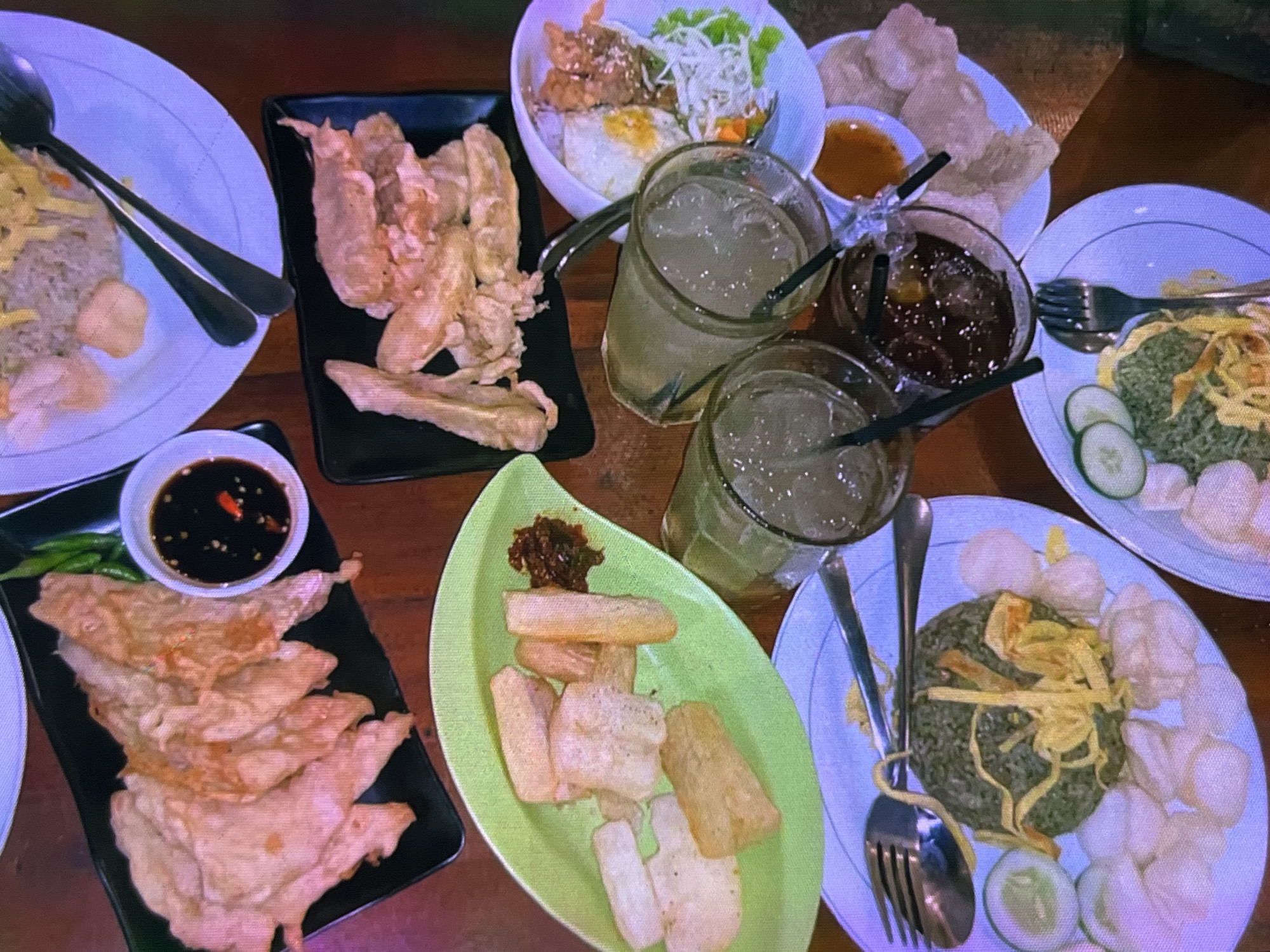 The image depicts a top-down view of a table laden with Indonesian food, likely at a casual eatery in Cadek, Indonesia. Several plates of fried items, including what appears to be tempeh and possibly other vegetables, are arranged on black rectangular platters. One platter features fried food with a small dish of dark dipping sauce containing chili. Another green leaf-shaped plate holds pieces of fried cassava or a similar starch, accompanied by a dollop of chili paste.
Two glasses filled with ice and a clear beverage, likely iced tea or soda, are visible, each with a straw. A third glass contains a darker, bubbly liquid, also with a straw. Adjacent to these drinks, on a white plate, is a portion of fried rice topped with shredded egg, accompanied by lettuce and a fried egg. Another white plate holds a portion of green-colored rice, garnished with shredded egg, cucumber slices, and an assortment of crackers or crisps. A third white plate, partially visible, appears to hold a similar arrangement of food. Cutlery, including forks and spoons, is placed on some of the plates. The food is set on a wooden table, suggesting an informal dining setting. The lighting appears to be ambient, casting a warm tone over the scene.