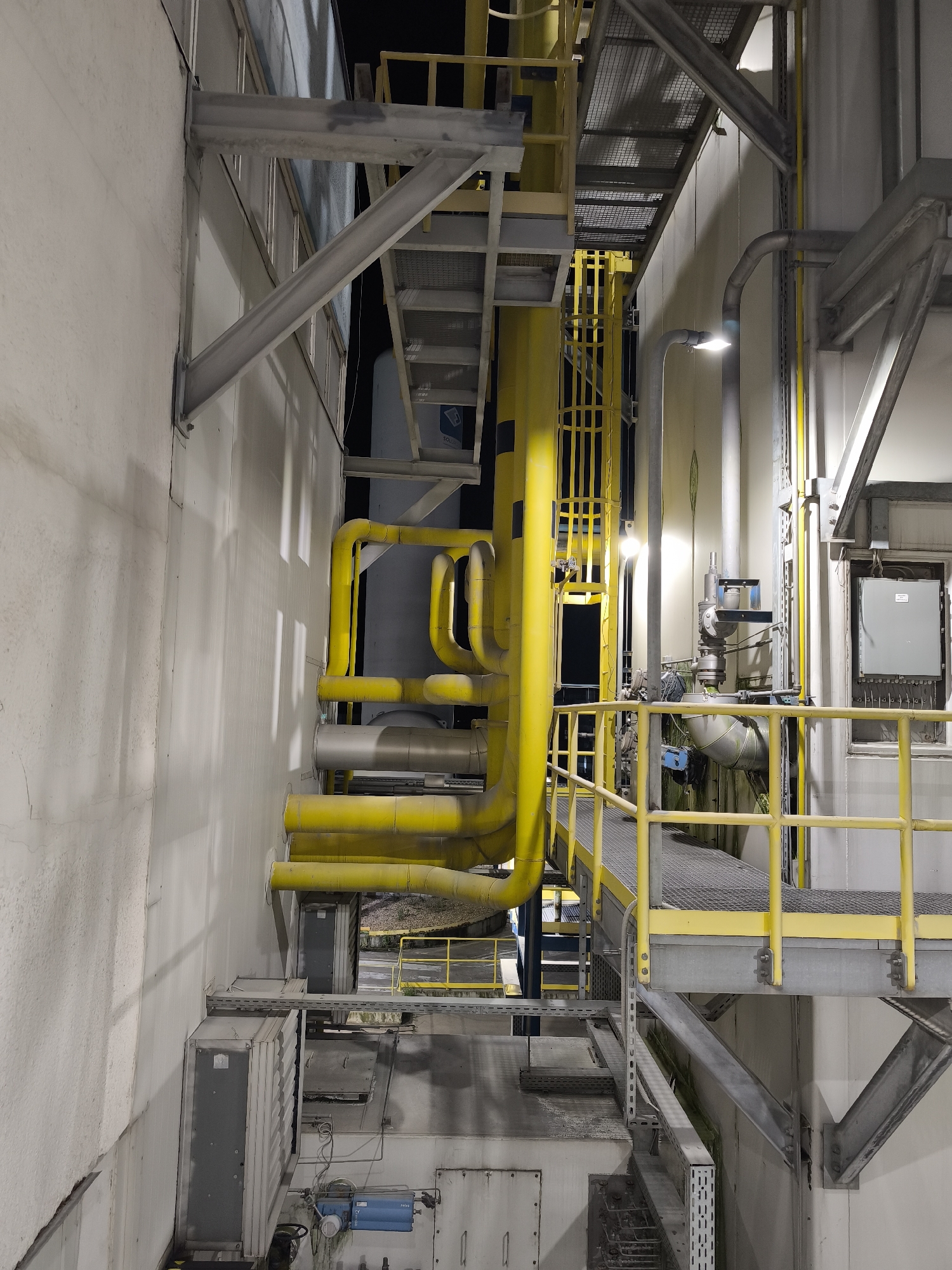 The image depicts an industrial facility with a complex network of yellow pipes and metal structures. The scene is dimly lit, with artificial lights illuminating certain areas. In the foreground, a white wall is visible, with visible shadows cast by the pipes and overhead structures. Several large, rectangular gray electrical boxes are mounted on the wall at different levels. A blue filtration unit is also visible near the bottom of the frame.
Dominating the midground is a series of large diameter yellow pipes that snake across the scene, supported by metal structures. Some of these pipes are insulated with gray material. A metal walkway with a yellow railing runs through the midground, providing access to different levels of the facility. Several metal staircases with grated treads are also visible, further emphasizing the industrial nature of the location.
In the background, more pipes and industrial equipment are present, along with the dark exterior of buildings. The overall impression is one of a functional, heavily industrialized area, likely involved in manufacturing or processing. The image appears to be taken at night or in a poorly lit indoor environment. There are no people or visible signs of activity. The specific location is not discernible from the image itself.