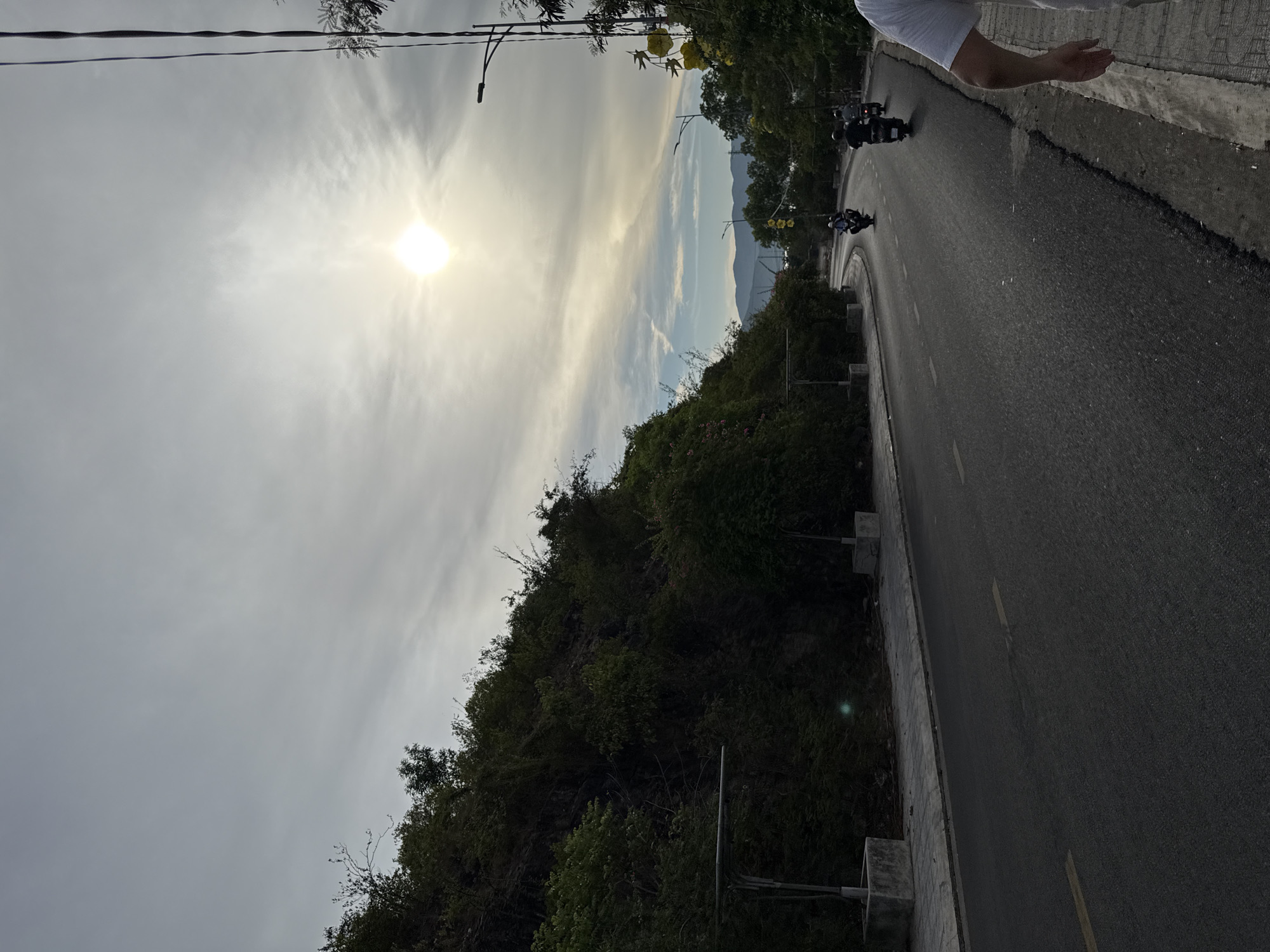 The image depicts a coastal road in Nha Trang, Vietnam, under a cloudy sky with the sun shining brightly. The road curves to the right, flanked by a lush, green hillside on the left and a sidewalk and more vegetation on the right. Several motorbikes are visible on the road, with riders wearing helmets. A person's arm and shoulder are visible on the far right edge of the frame, suggesting they are walking on the sidewalk. The environment appears to be a scenic route, possibly on the outskirts of the city, with mountains visible in the distance. The overall atmosphere is that of a typical day in a tropical coastal city.