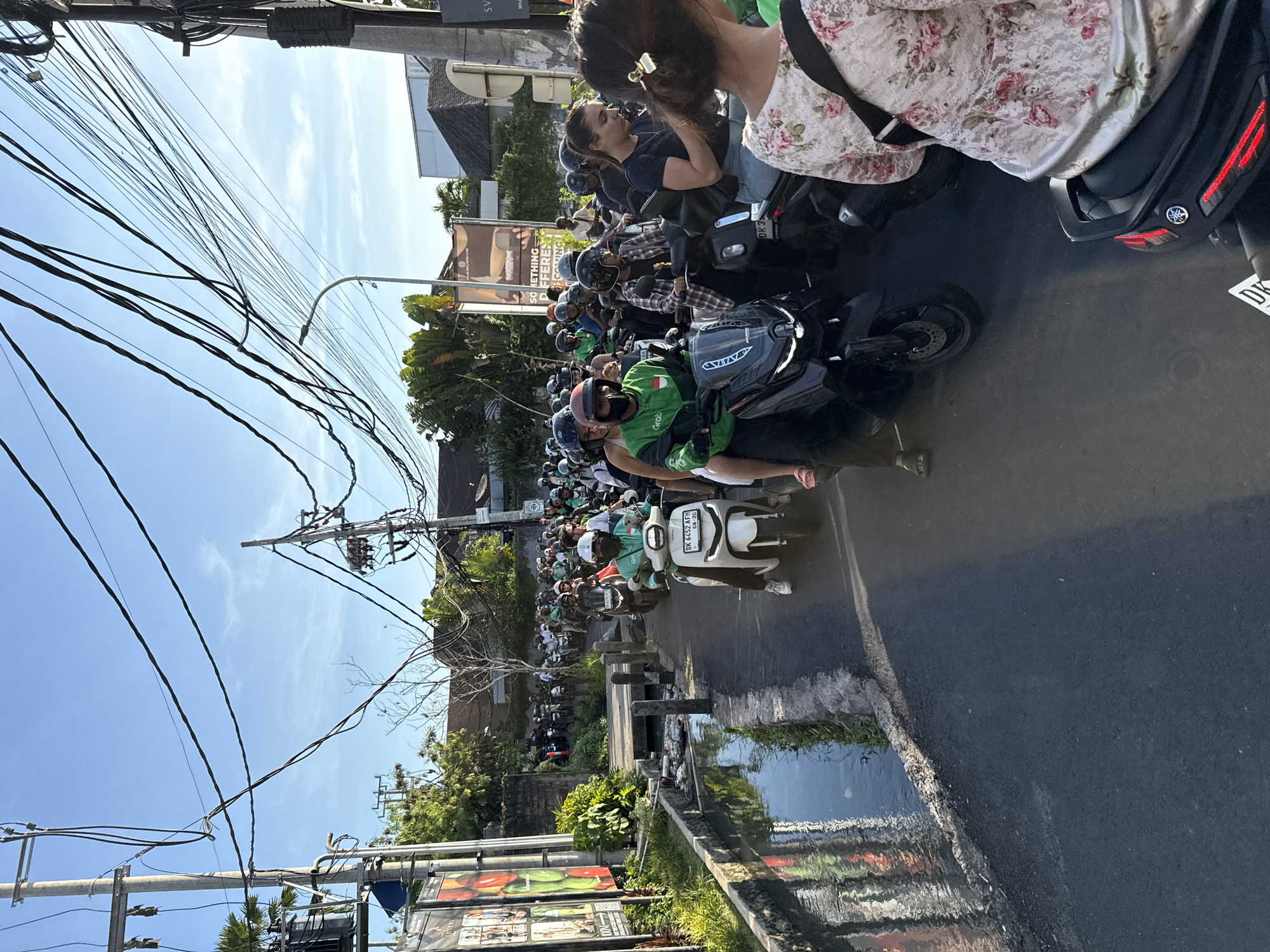 The image depicts a congested street in North Kuta, Indonesia, filled with a large number of motorcycles and scooters. Most drivers wear helmets. Many motorcyclists wear a green jacket with "Grab" displayed on it. Overhead, a dense network of electrical wires is visible. A person in a floral shirt stands in the foreground. A water-filled drainage ditch runs alongside the road. A billboard showing a group of individuals in different poses stands alongside the street. Buildings are visible in the background. The sky is clear and blue.