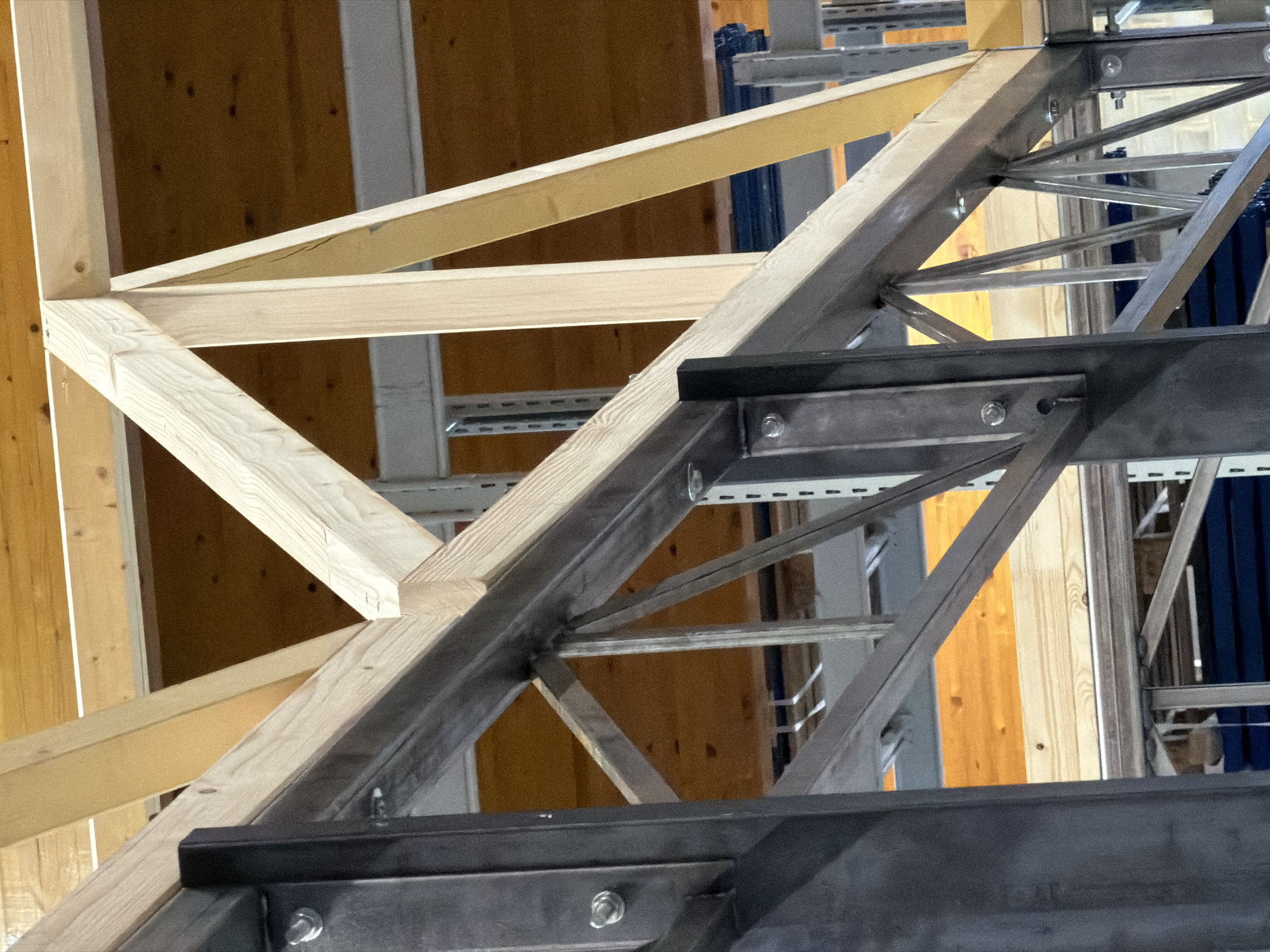 The image shows a close-up view of an industrial storage facility in Leudelange, Luxembourg. The primary subjects are structural elements, including dark grey steel beams and light-colored wooden beams forming a complex framework. The steel beams appear to be part of a shelving or racking system, with visible bolts and connections. The wooden beams are arranged in a truss-like structure, likely supporting a roof or upper level. In the background, glimpses of other shelving units and a warm, orange-toned wooden surface can be seen, possibly the floor or ceiling of a different level. The lighting suggests an indoor environment with some ambient light, creating shadows and highlighting the textures of the materials. There are no people or active processes visible in the image.