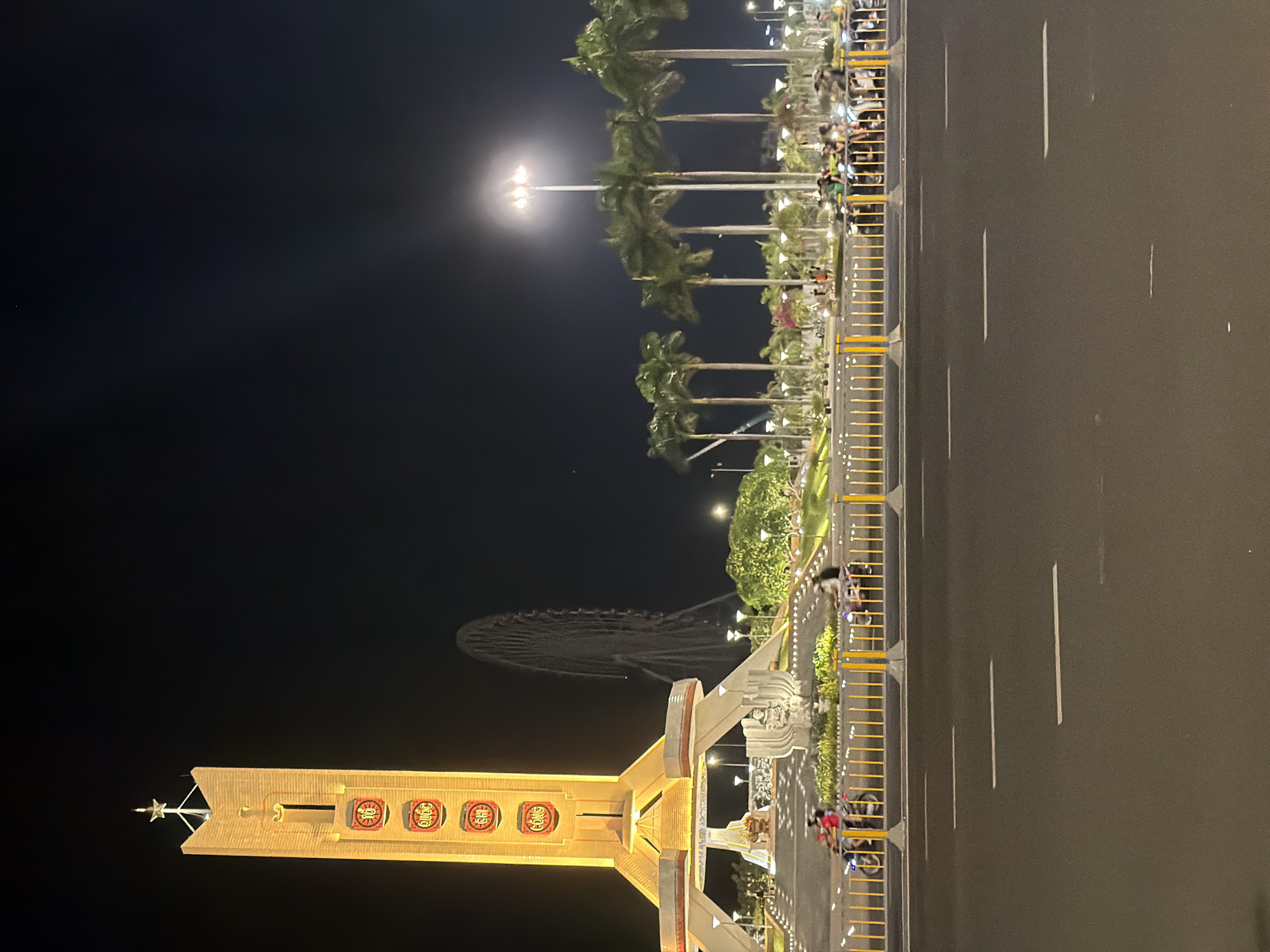 This is a nighttime photograph of a city square in Da Nang, Vietnam. The most prominent feature is a tall, illuminated monument with a star at its apex, decorated with circular emblems containing Vietnamese characters. To the right of the monument, a large Ferris wheel is visible in the background, partially obscured by palm trees. The foreground shows a road with lane markings, and a yellow barrier separates the road from a planted area. Several motorbikes are parked or being ridden behind the barrier, with some riders visible. The environment is illuminated by streetlights and the monument itself, creating a slightly hazy glow in the dark sky.